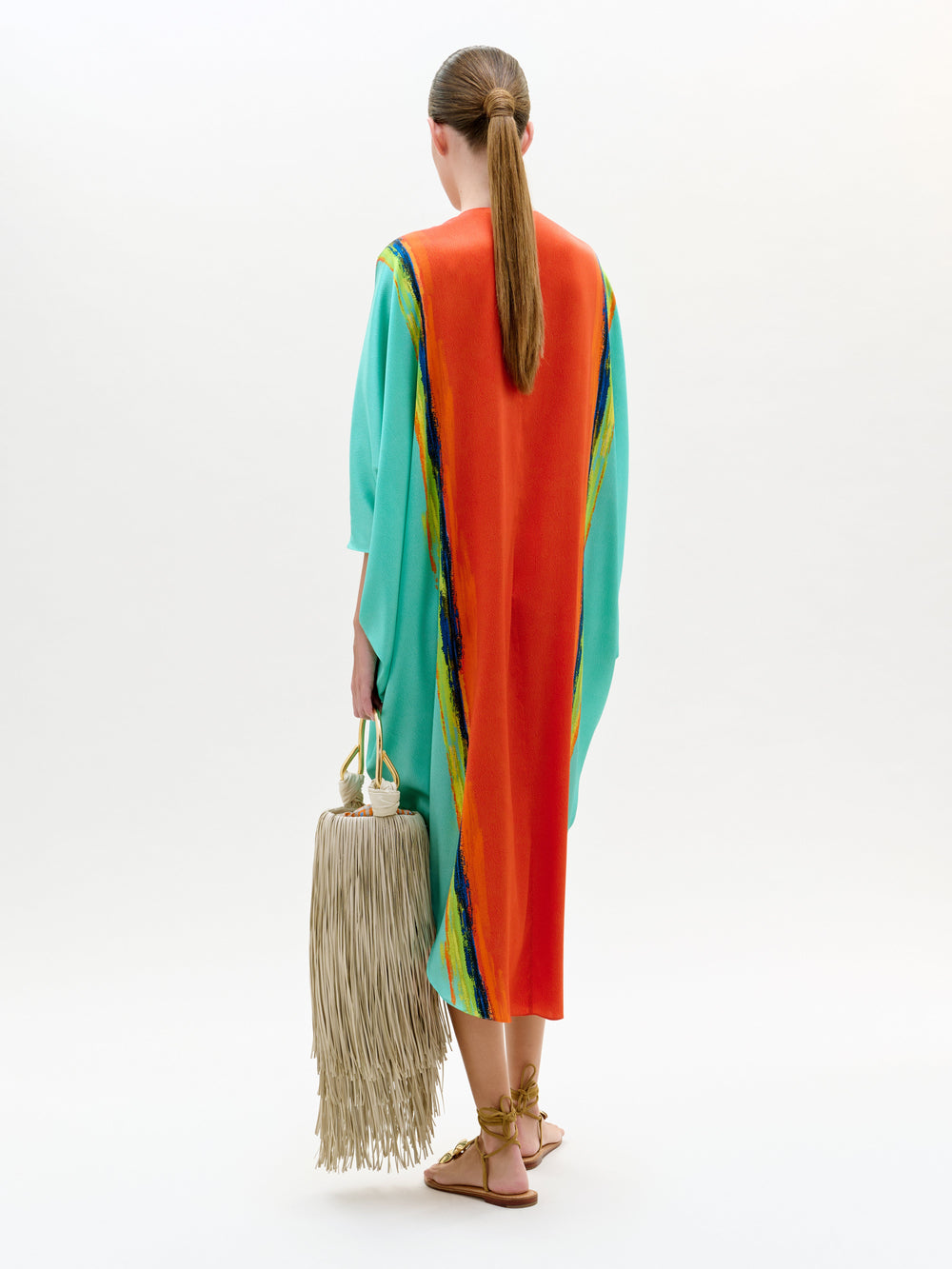 Una persona, de espaldas, lleva la túnica Nidia, un maxi abstracto turquesa y naranja confeccionado con tela italiana en Colombia. Lleva un bolso grande con flecos, sandalias color canela y una coleta baja sobre un fondo blanco liso.