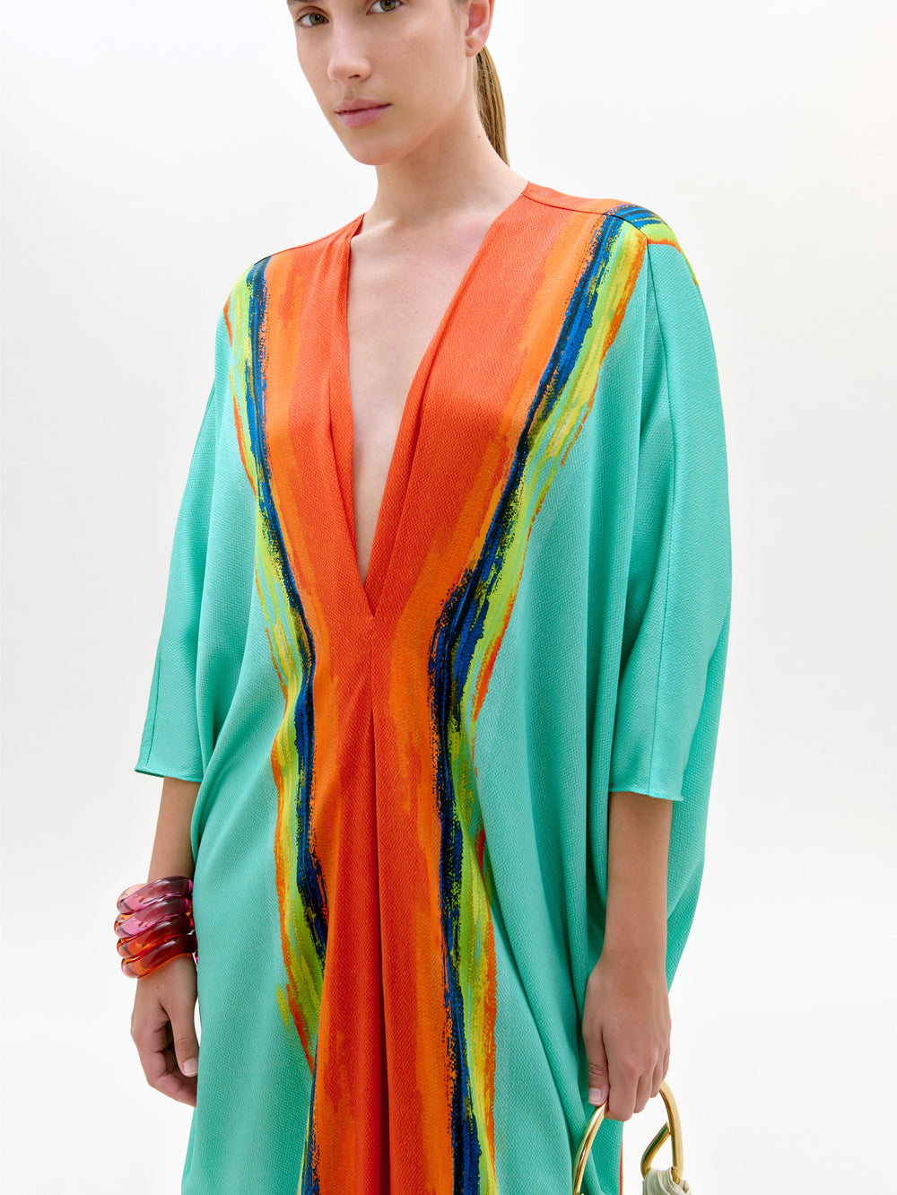 Con la túnica Nidia -una maxi suelta de color aguamarina con mangas anchas y atrevidas rayas verticales naranjas y multicolores en tejido italiano-, una persona aparece sobre un fondo liso, con un bolso dorado y gruesas pulseras rojas.