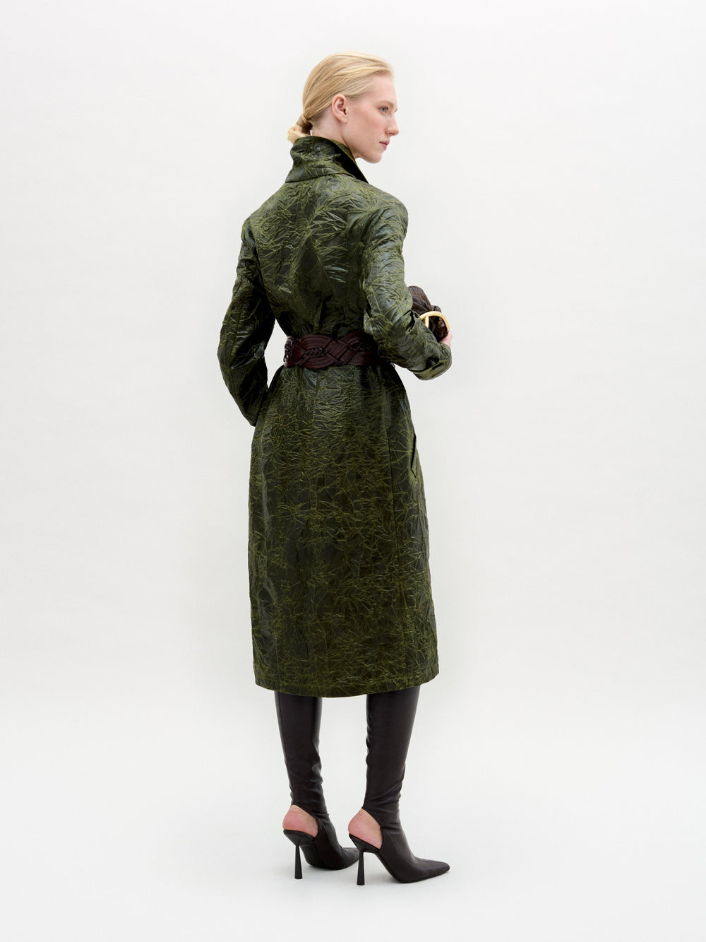 Una persona, de espaldas, lleva el Nilo Trench -un abrigo verde texturizado fabricado en Colombia- ceñido a la cintura, combinado con botas negras de tacón y guantes marrón oscuro. El fondo es liso y de color claro.