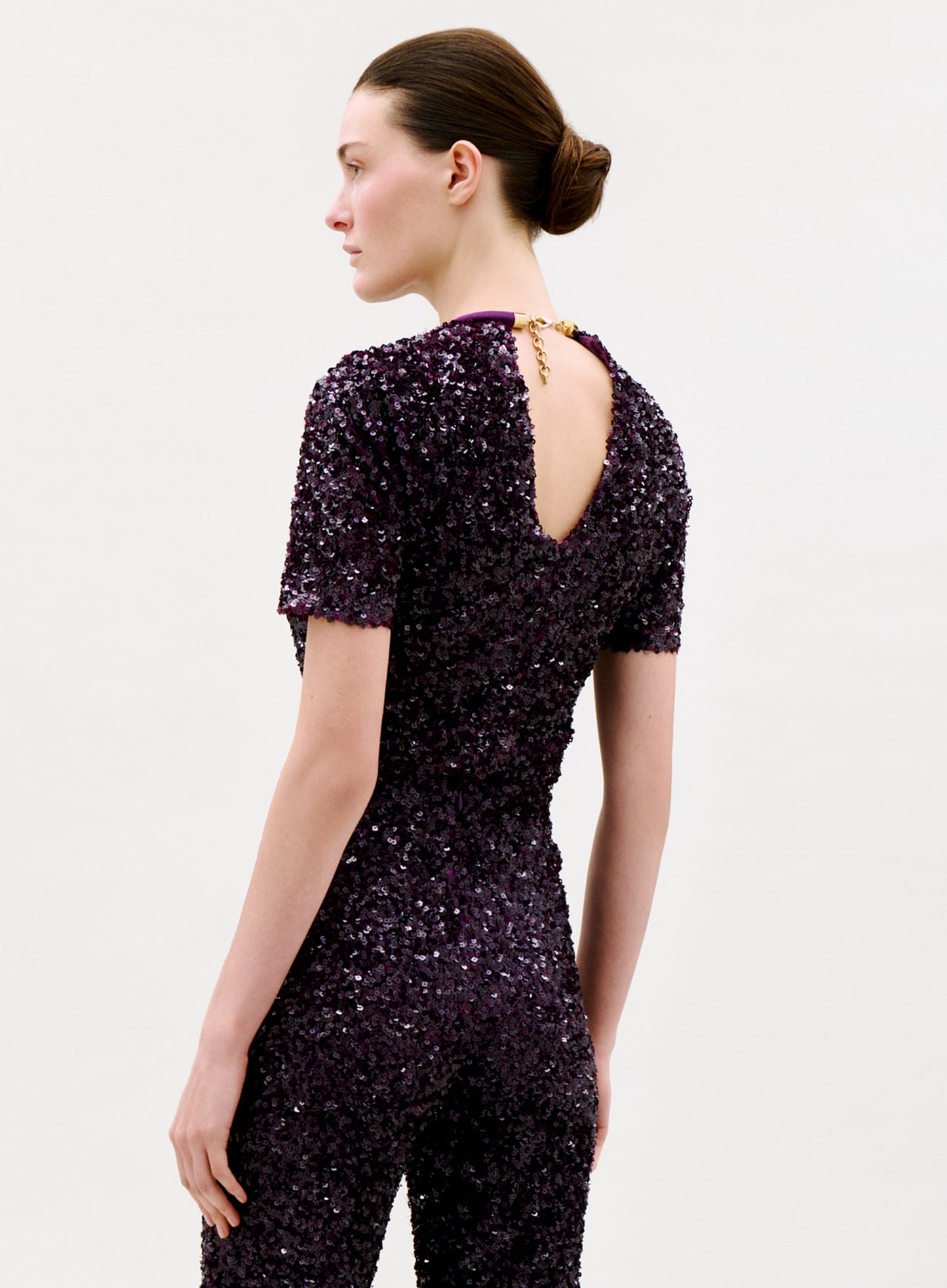 A woman with brown hair in a bun wears the Napoles Blouse, a dark purple sequin top with gold chain detail, standing against a plain light background and looking down over her shoulder.
