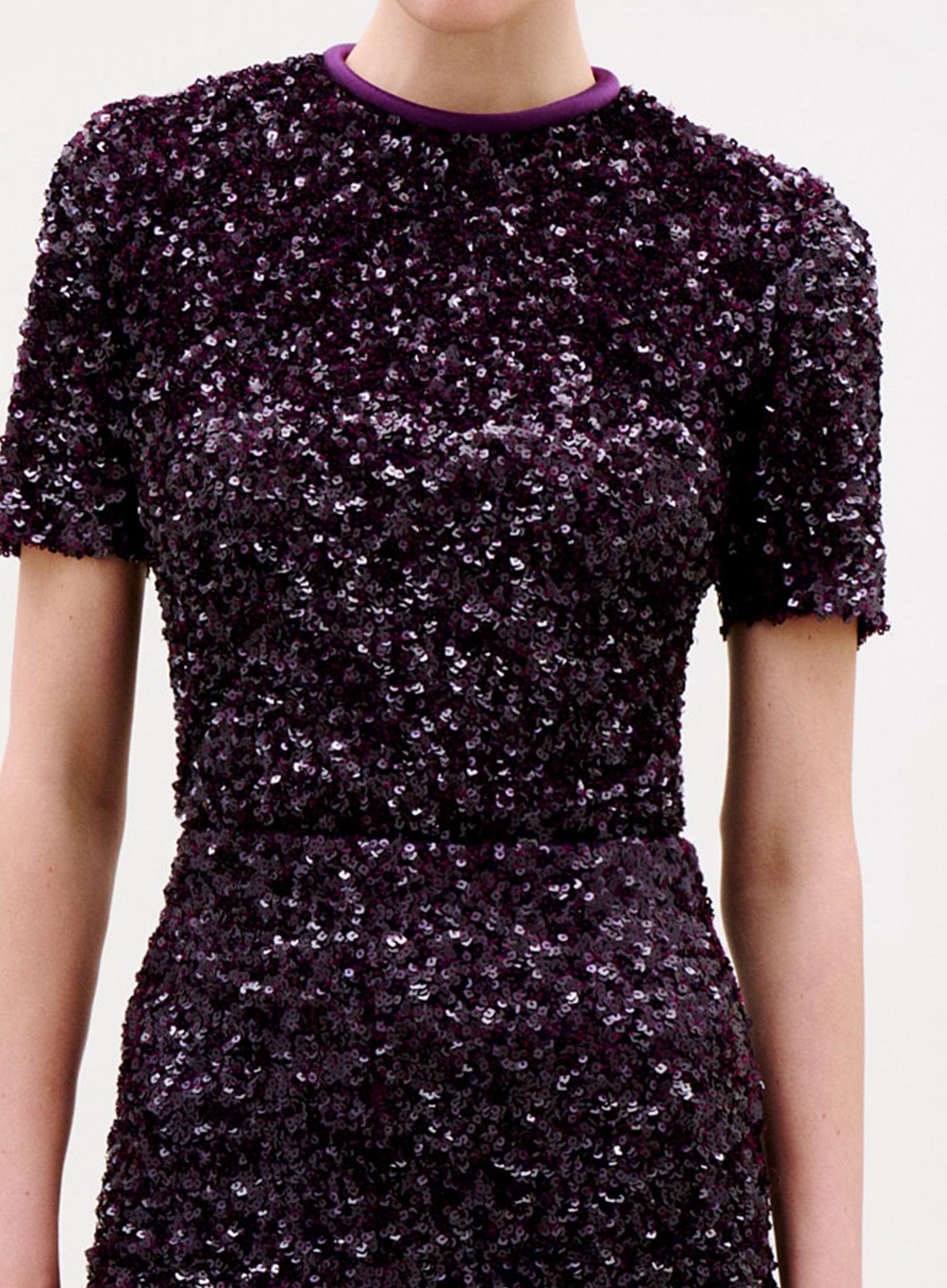 A woman with brown hair in a bun wears the Napoles Blouse, a dark purple sequin top with gold chain detail, standing against a plain light background and looking down over her shoulder.