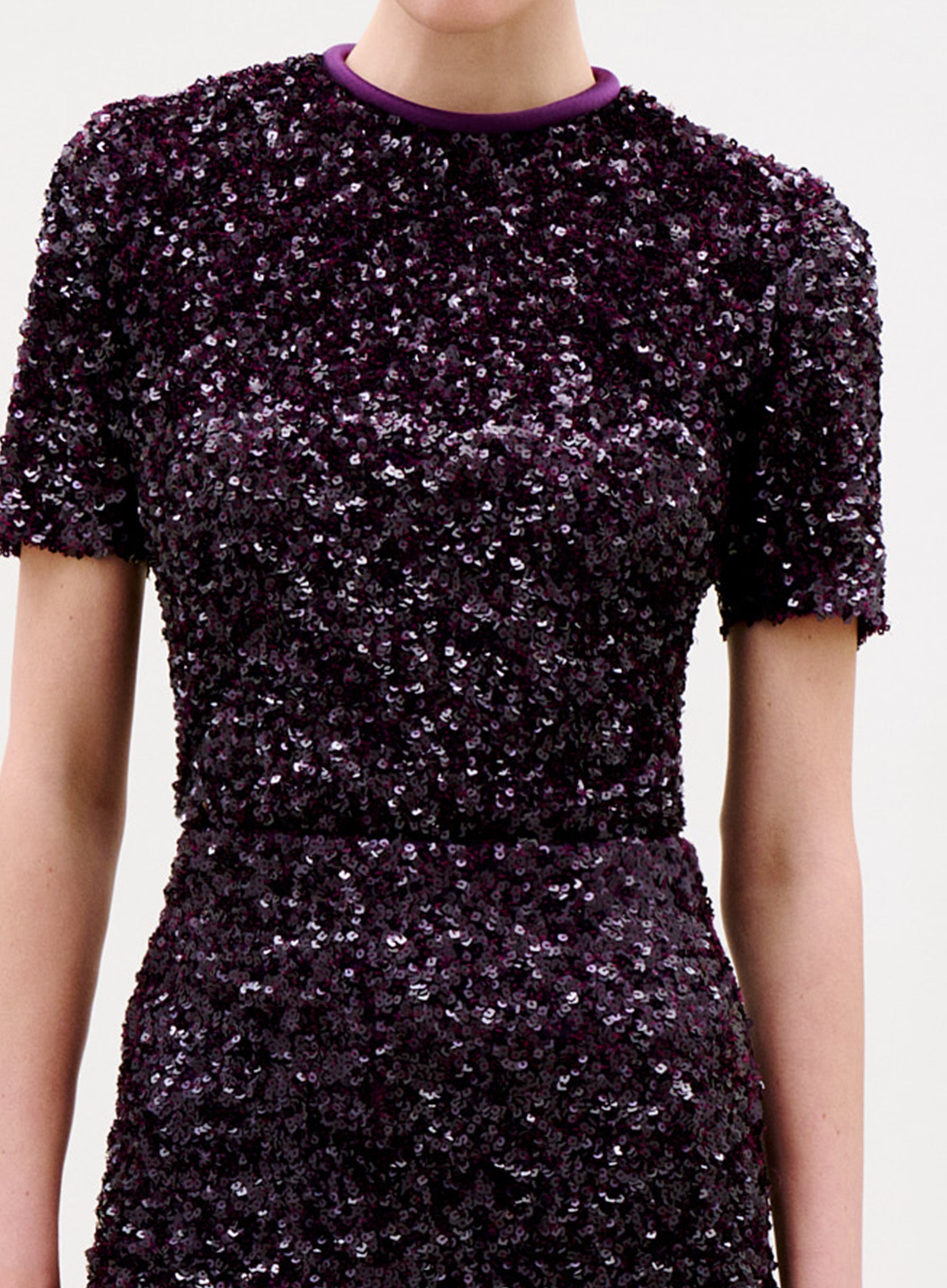 A woman with brown hair in a bun wears the Napoles Blouse, a dark purple sequin top with gold chain detail, standing against a plain light background and looking down over her shoulder.