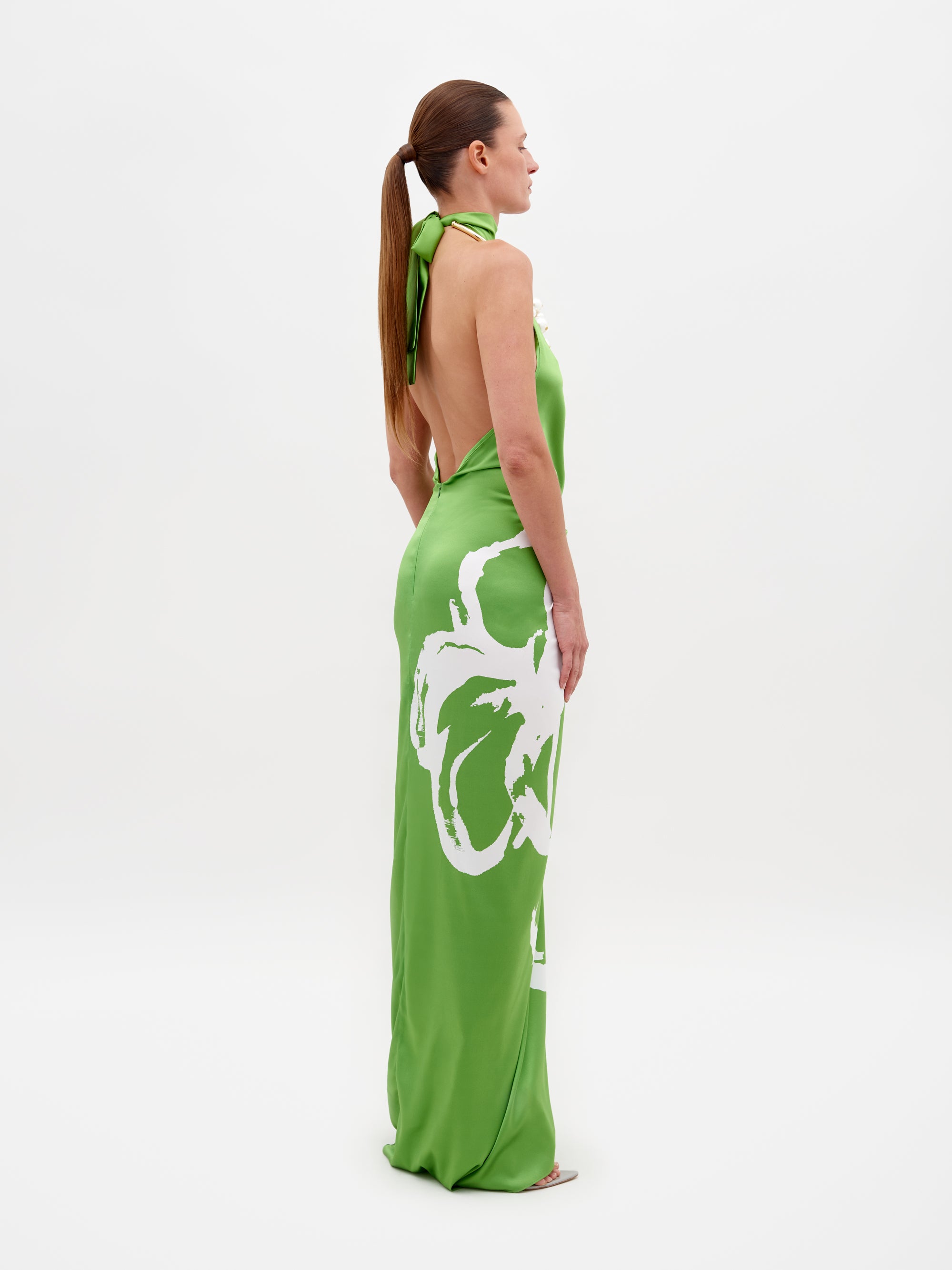 Una mujer de perfil aparece de pie frente a un fondo sencillo, luciendo el Vestido Olatz, un vestido largo verde con la espalda descubierta. Tiene un cuello alto y un gran diseño floral blanco en el lateral. Su cabello largo está peinado en una cola de caballo.