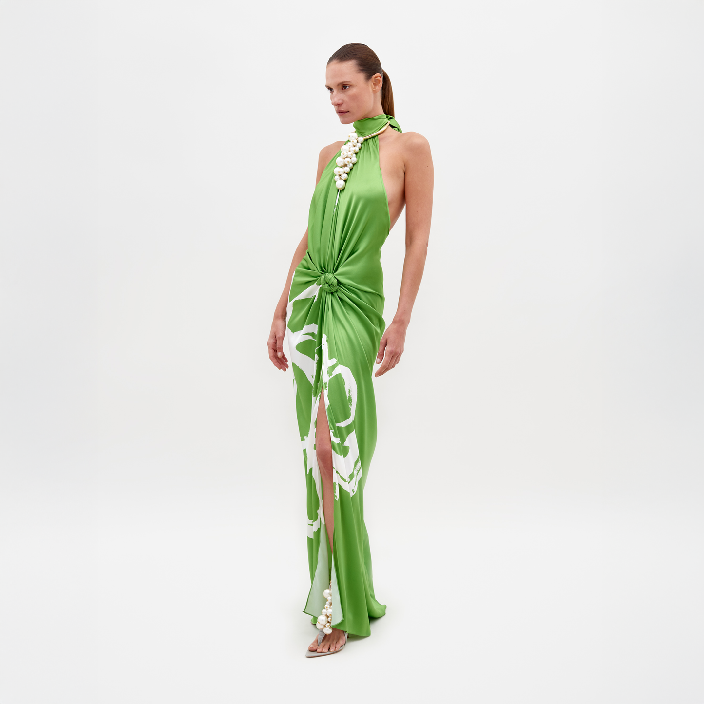 Sobre un fondo blanco, una mujer lleva el Vestido Olatz, un vestido largo sin mangas de seda verde brillante con un estampado floral abstracto. Presenta un diseño de cuello halter, abertura alta y detalle de cintura fruncida. El look se completa con sandalias de tacón alto y cabello largo recogido.
