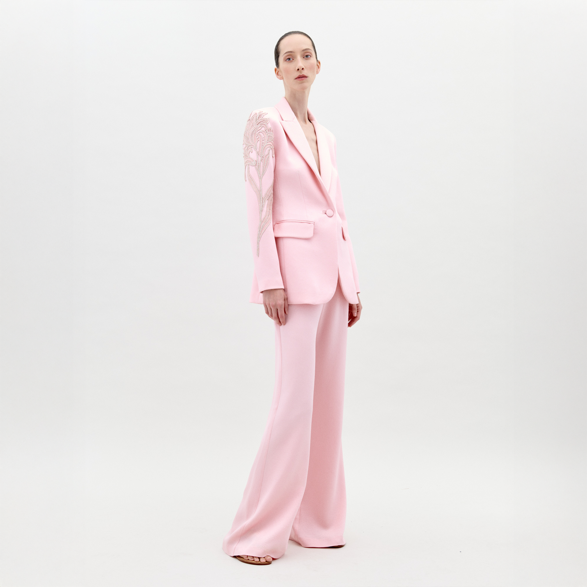 Una persona está de pie sobre un fondo blanco liso, vistiendo el Pantalón Palermo (pantalones de tiro alto y pierna ancha de satén martillado rosa claro) y un blazer a juego con un diseño floral en un hombro. Con el cabello recogido, mira hacia adelante.
