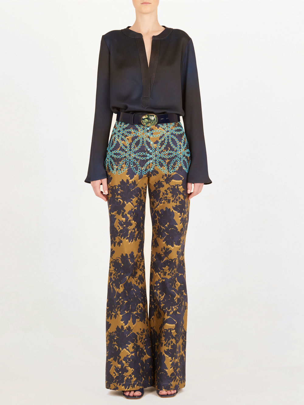 Una persona de pie, vestida con una blusa de algodón de manga larga y el pantalón de cintura alta Como Camo Navy con motivos de flor de filigrana en marrón, azul marino y verde azulado, rematado con un cinturón negro con hebilla decorativa.