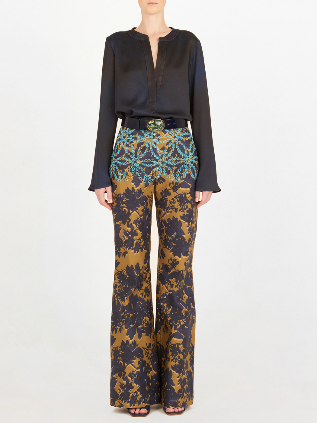 Una persona de pie, vestida con una blusa de algodón de manga larga y el pantalón de cintura alta Como Camo Navy con motivos de flor de filigrana en marrón, azul marino y verde azulado, rematado con un cinturón negro con hebilla decorativa.