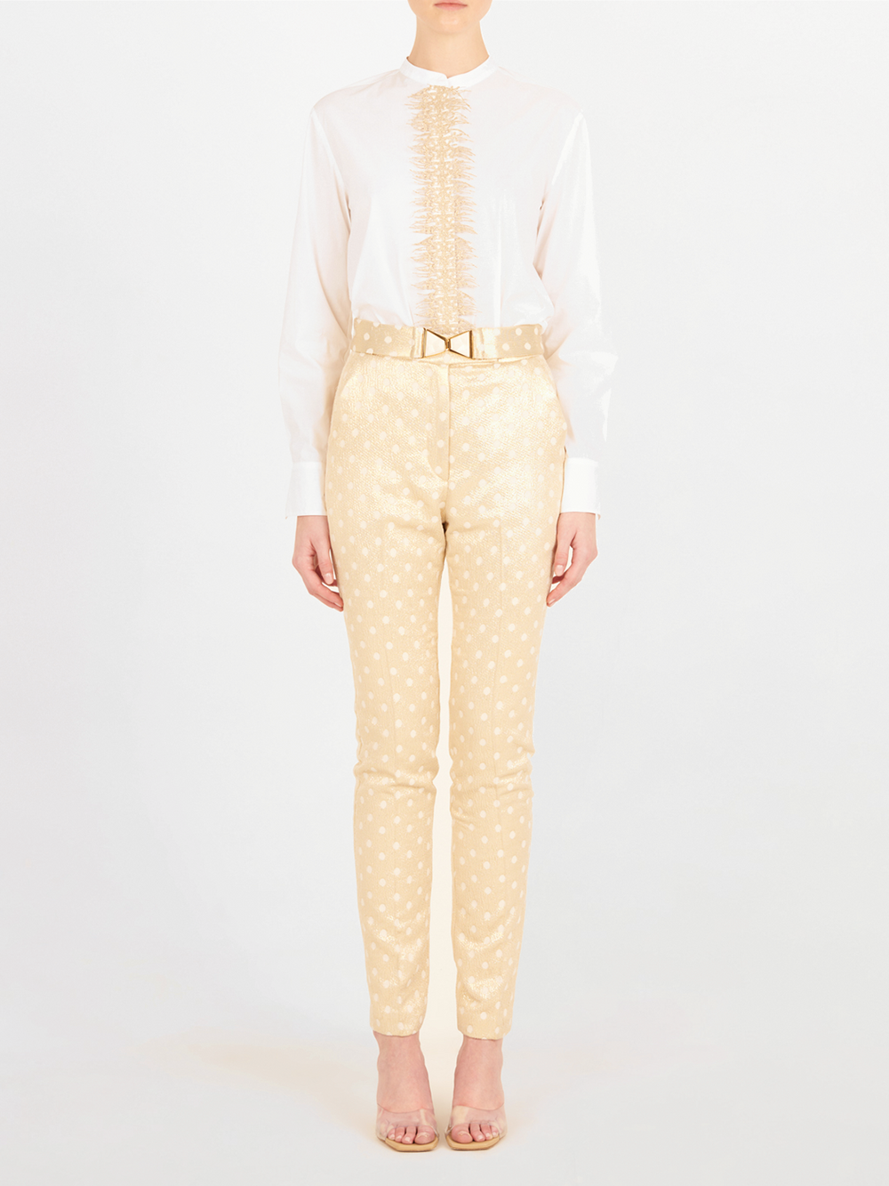 Una persona de pie sobre un fondo blanco, con una blusa blanca de manga larga con volantes, el Pantalón Orion Gold Inverted en beige con un cinturón estampado, y tacones beige abiertos.