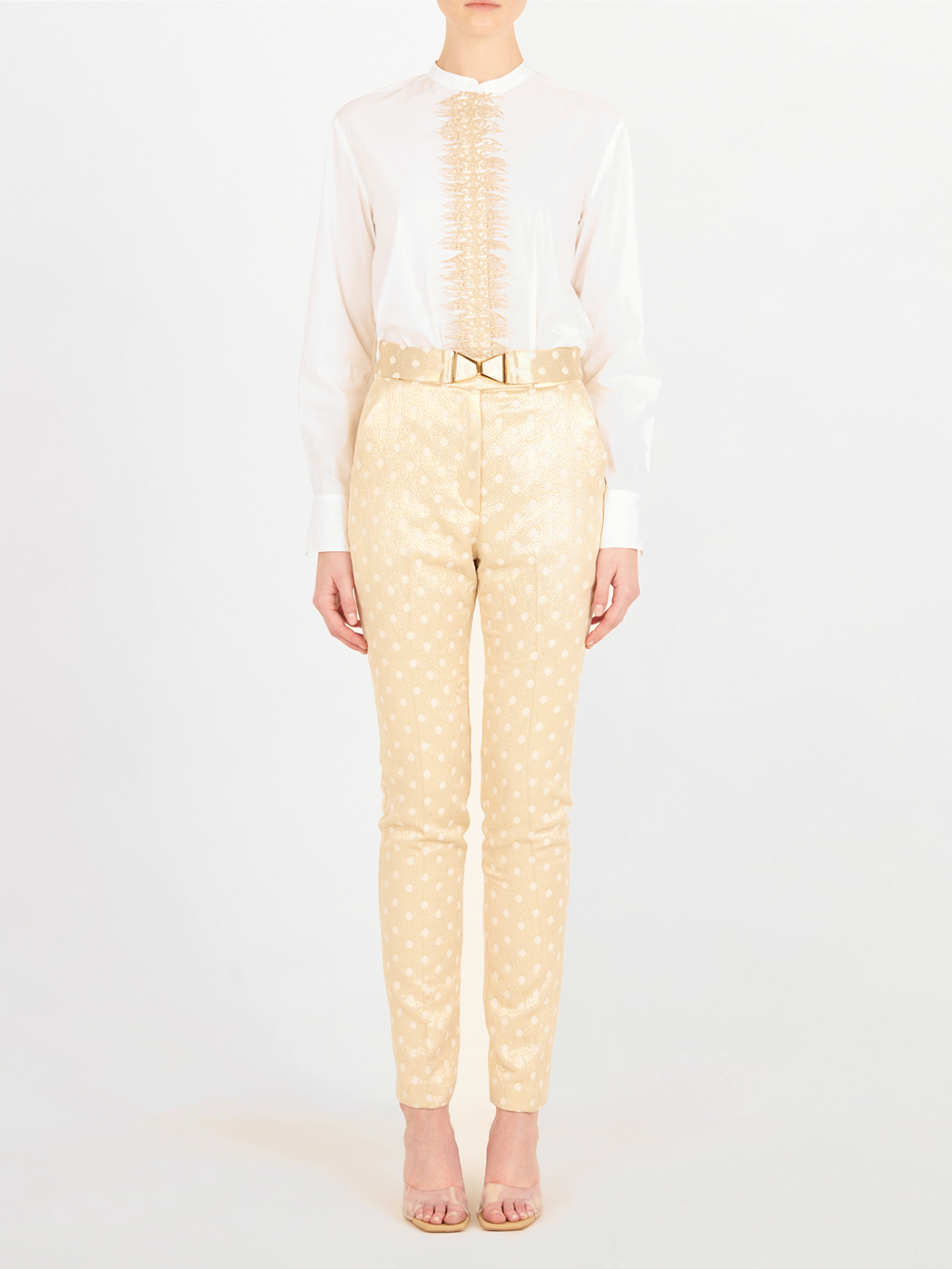 Una persona de pie sobre un fondo blanco, con una blusa blanca de manga larga con volantes, el Pantalón Orion Gold Inverted en beige con un cinturón estampado, y tacones beige abiertos.