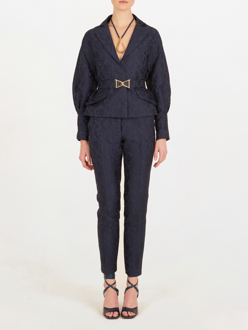 Una persona lleva el Pantalón Orion Navy, un traje pantalón de talle alto en azul marino oscuro con textura, cinturón en la cintura y escote profundo en V, combinado con sandalias de tacón de tiras negras, sobre un fondo blanco liso.