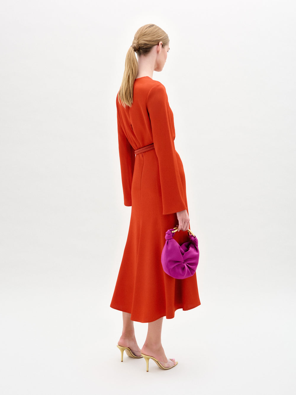 Una mujer con el pelo rubio recogido en una coleta baja lleva el vestido rojo Pesaro y sandalias de tacón beige, sosteniendo un bolso de color morado brillante sobre un fondo liso de color claro.