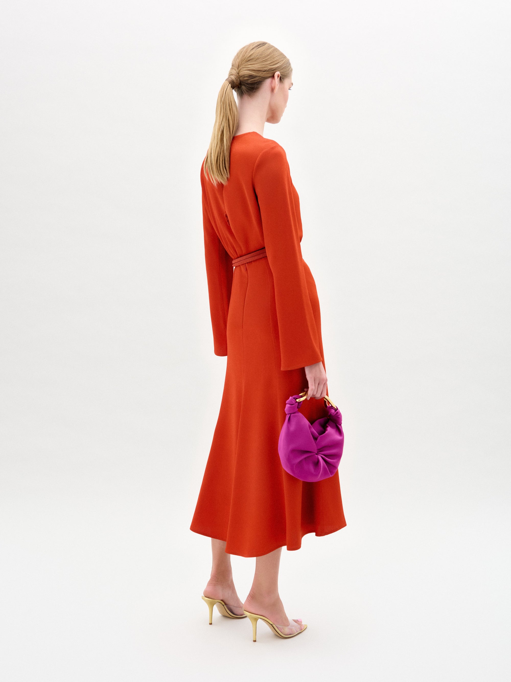 Una mujer con el pelo rubio recogido en una coleta baja lleva el vestido rojo Pesaro y sandalias de tacón beige, sosteniendo un bolso de color morado brillante sobre un fondo liso de color claro.