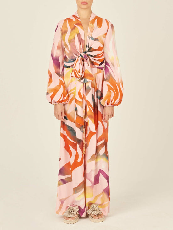 The Honey Blouse Blush Prismatic is a long-sleeve top with a vibrant abstract zebra print in pink, orange, yellow, and white. It features a front tie knot design and a flowy silhouette.