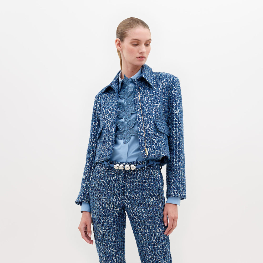 Una persona que viste la Chaqueta Perla y pantalones a juego se encuentra de pie sobre un fondo liso. El atuendo incluye una blusa celeste con volados y un cinturón blanco con elementos decorativos que realzan la silueta estructurada, mientras su cabeza está ligeramente girada hacia un lado.