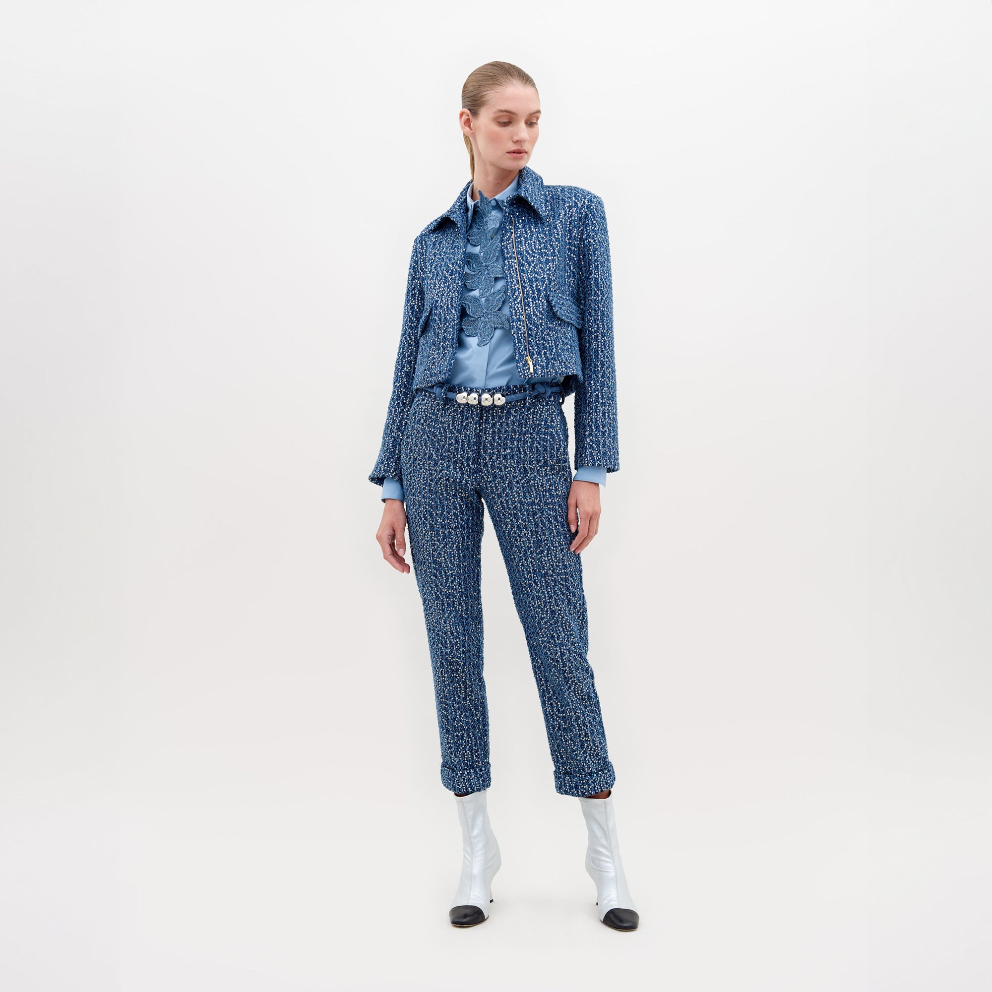 Una persona está de pie con un pantalón y una chaqueta estampados en azul hechos de la exquisita tela Textil Italiano, combinados con una camisa azul claro. Tiene el cabello de color claro atado hacia atrás y usa botas de tacón blancas con puntas negras sobre un fondo blanco liso.
