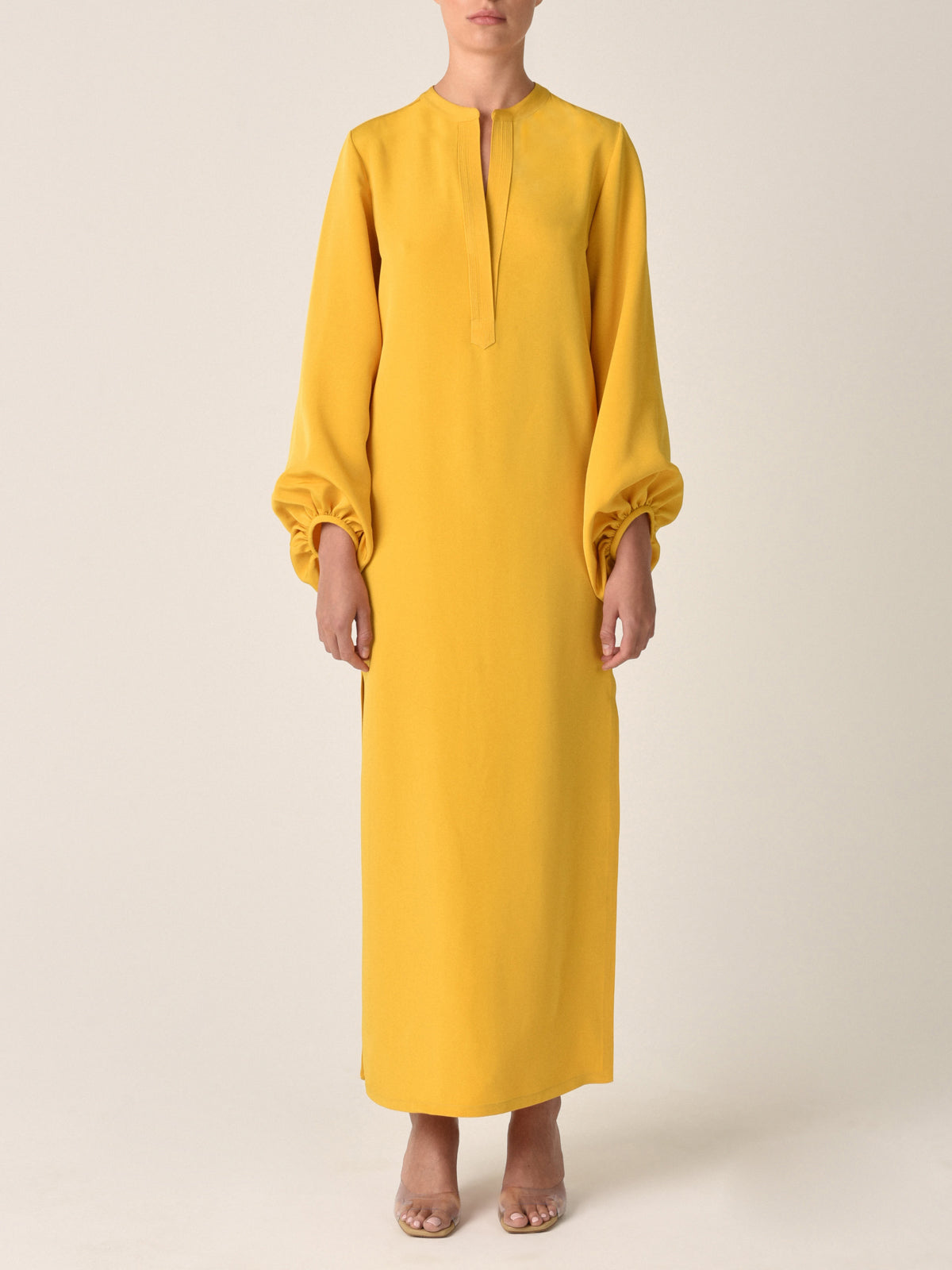 The Monferrato Tunic Amber is a long, mustard yellow dress with a high neckline and romantic balloon sleeves gathered at the wrists, featuring a small V-cut at the front. Perfect for Resort 2023, this amber-hued crepe tunic adds an elegant touch to your vacation wardrobe.