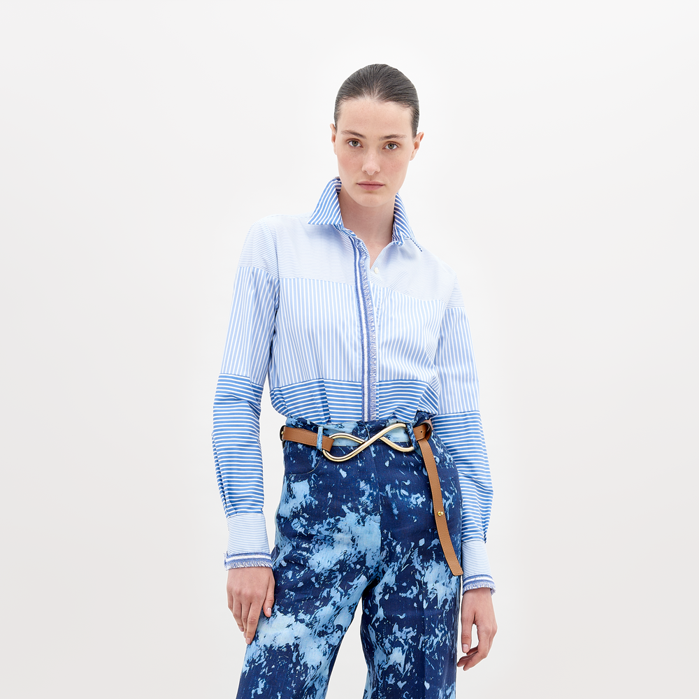 Vestido con una Camisa Raina, una camisa de manga larga 100% algodón azul claro con estampado de rayas, metida por dentro de unos pantalones azules con efecto tie-dye. El look se completa con un cinturón marrón con una gran hebilla de metal, todo ello sobre un fondo blanco liso.
