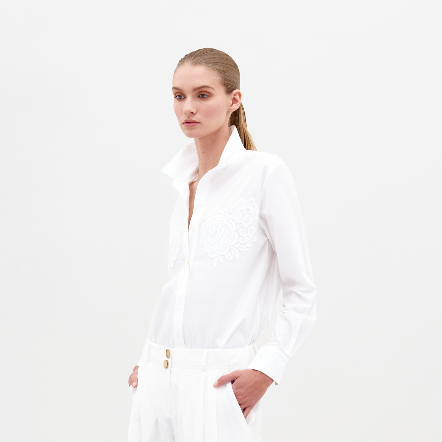 Una persona con el pelo largo atado hacia atrás lleva una blusa raizel adornada con apliques florales y pantalones blancos con botones dorados. Está de pie sobre un fondo claro y liso con las manos en los bolsillos.