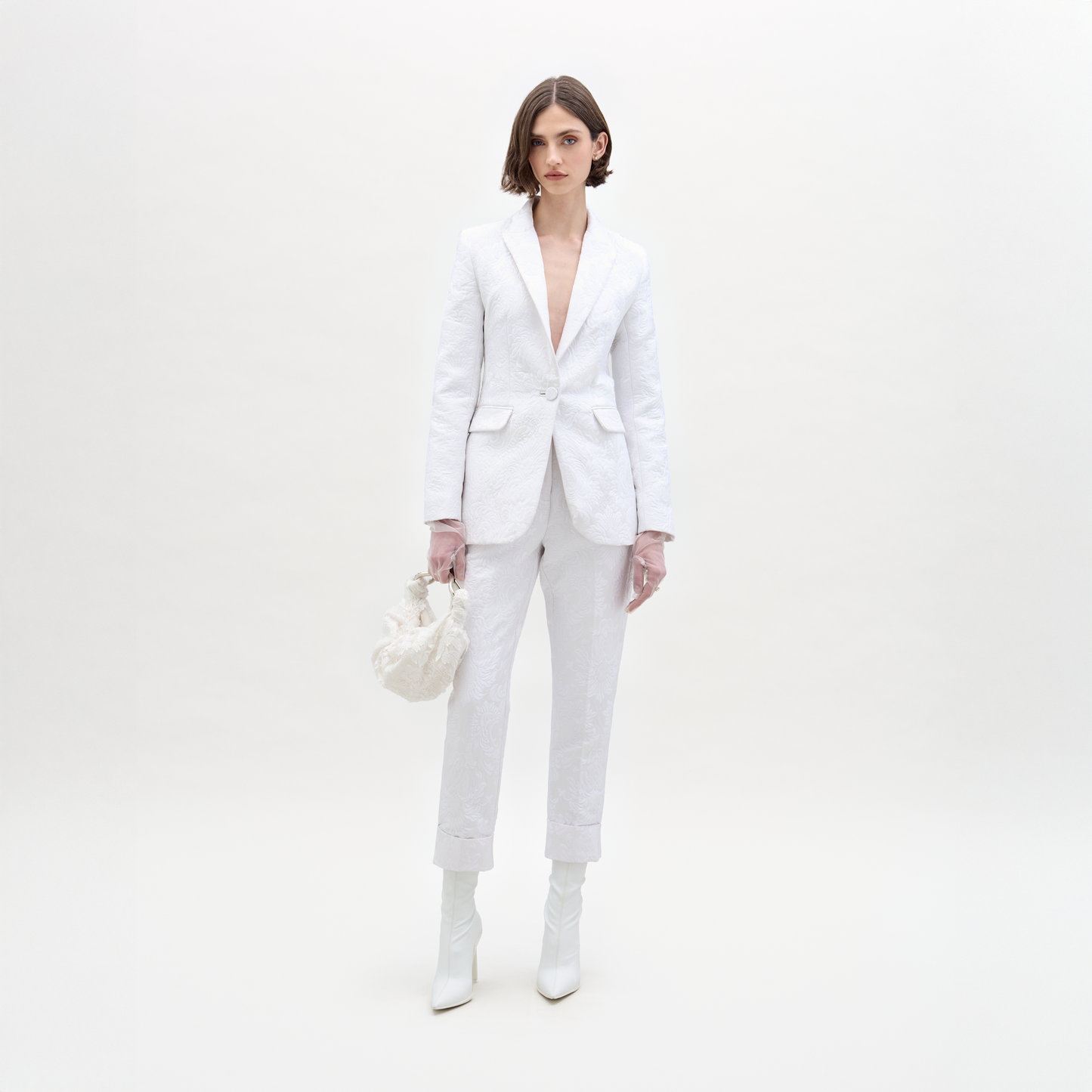 Una mujer de cabello castaño corto y expresión neutra está parada sobre un fondo blanco vistiendo la Chaqueta Rebeca, pantalones blancos, botines blancos y sosteniendo un pequeño bolso blanco.