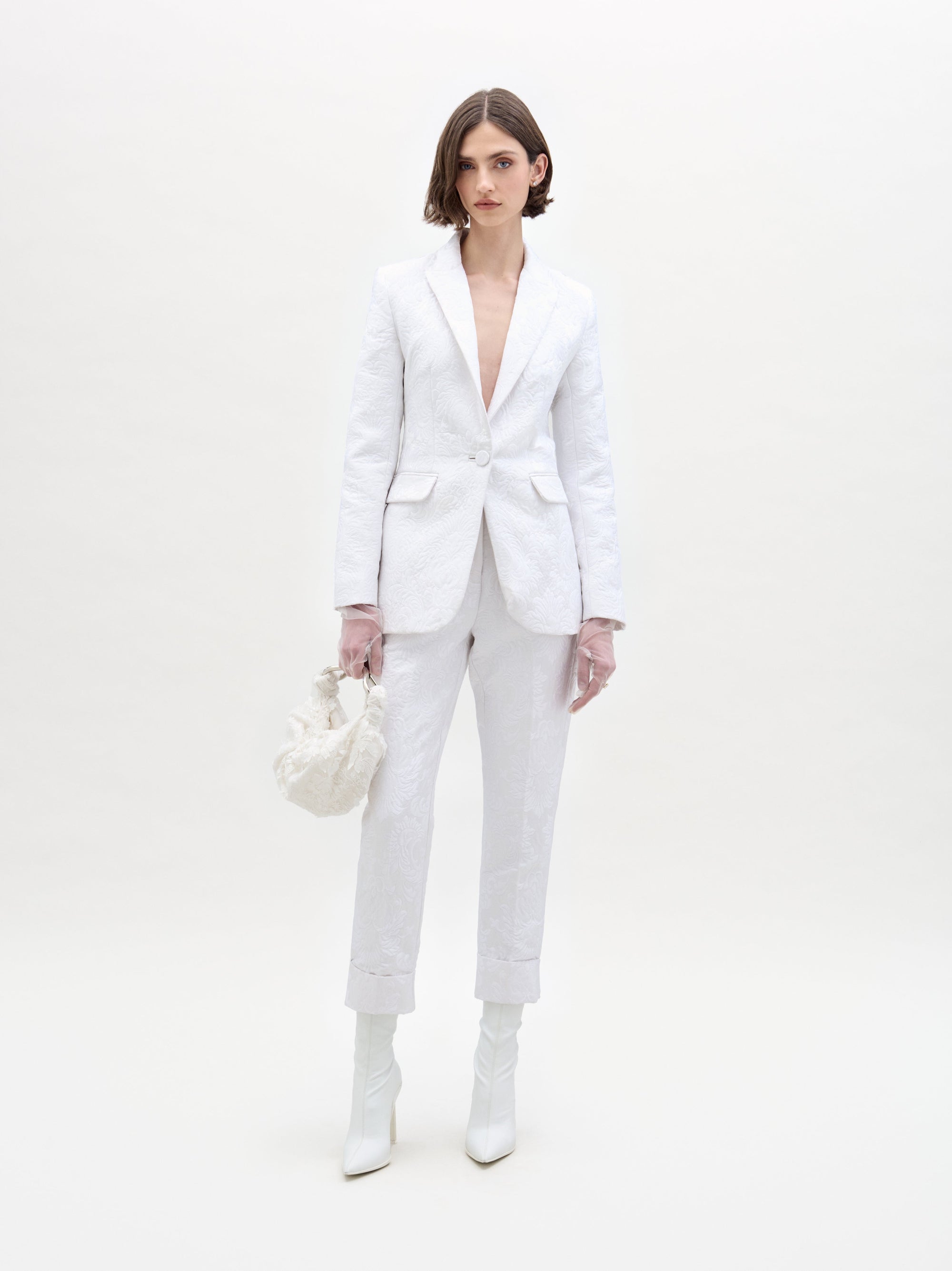 Una persona posa sobre un fondo sencillo, vestida con el pantalón Moad y una chaqueta blanca a juego. Lleva un bolso blanco, botas blancas y guantes transparentes.
