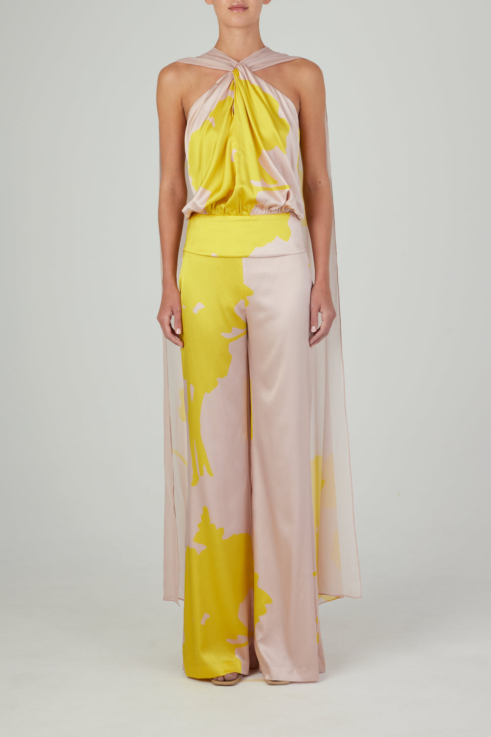 Luciendo la blusa Vicenta Amarillo Nude (un top halter sin mangas, rosa pálido y amarillo, con espalda drapeada) combinada con pantalones anchos con estampado abstracto sobre un fondo claro. Listo para envío en pocos días tras su pedido.