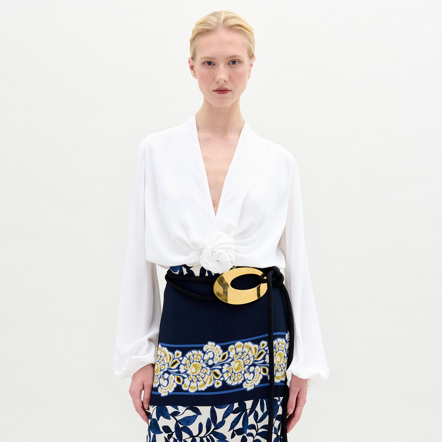 Una persona con cabello rubio recogido lleva la Blusa Rocío, una blusa blanca de manga larga con cuello en V y un gran detalle floral blanco en la cintura, combinada con un cinturón negro y dorado y una falda azul marino con flores amarillas y blancas. Hecha en Colombia.