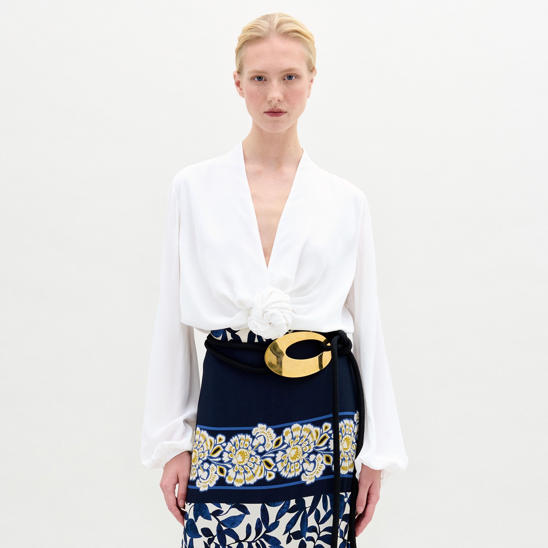 Una persona con cabello rubio recogido lleva la Blusa Rocío, una blusa blanca de manga larga con cuello en V y un gran detalle floral blanco en la cintura, combinada con un cinturón negro y dorado y una falda azul marino con flores amarillas y blancas. Hecha en Colombia.