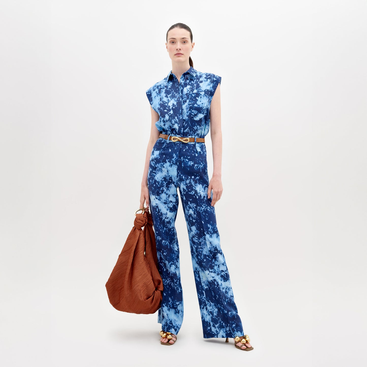 Una mujer se encuentra de pie sobre un fondo liso con el Pantalón Emine en azul tie-dye, 100% lino, sin mangas, con un cinturón marrón, sandalias doradas y sosteniendo un gran bolso marrón. Lleva el cabello liso recogido y una expresión neutra.