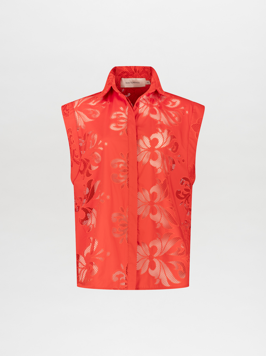 A person stands wearing a vibrant orange sleeveless top and matching pants with floral patterns, accessorized with a brown belt and holding a brown handbag. Pre-order the Rina Blouse Rouge Embroidery now to ensure it ships by the date of November 15th, 2024.