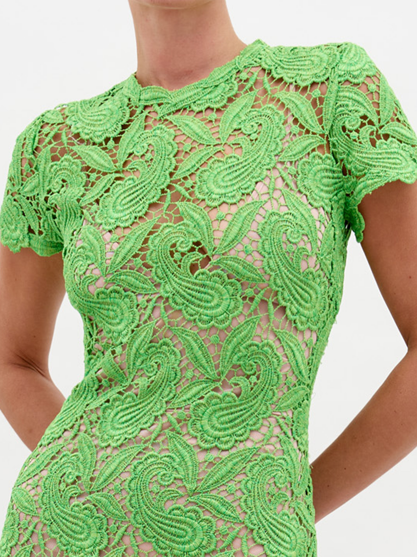 A person stands against a plain white background, wearing the Roxana Dress Green Guipure—a long, fitted lace dress with short sleeves. Hands are clasped behind the back. This elegant dress is available for pre-order now, with a scheduled ship date of February 15th, 2025.