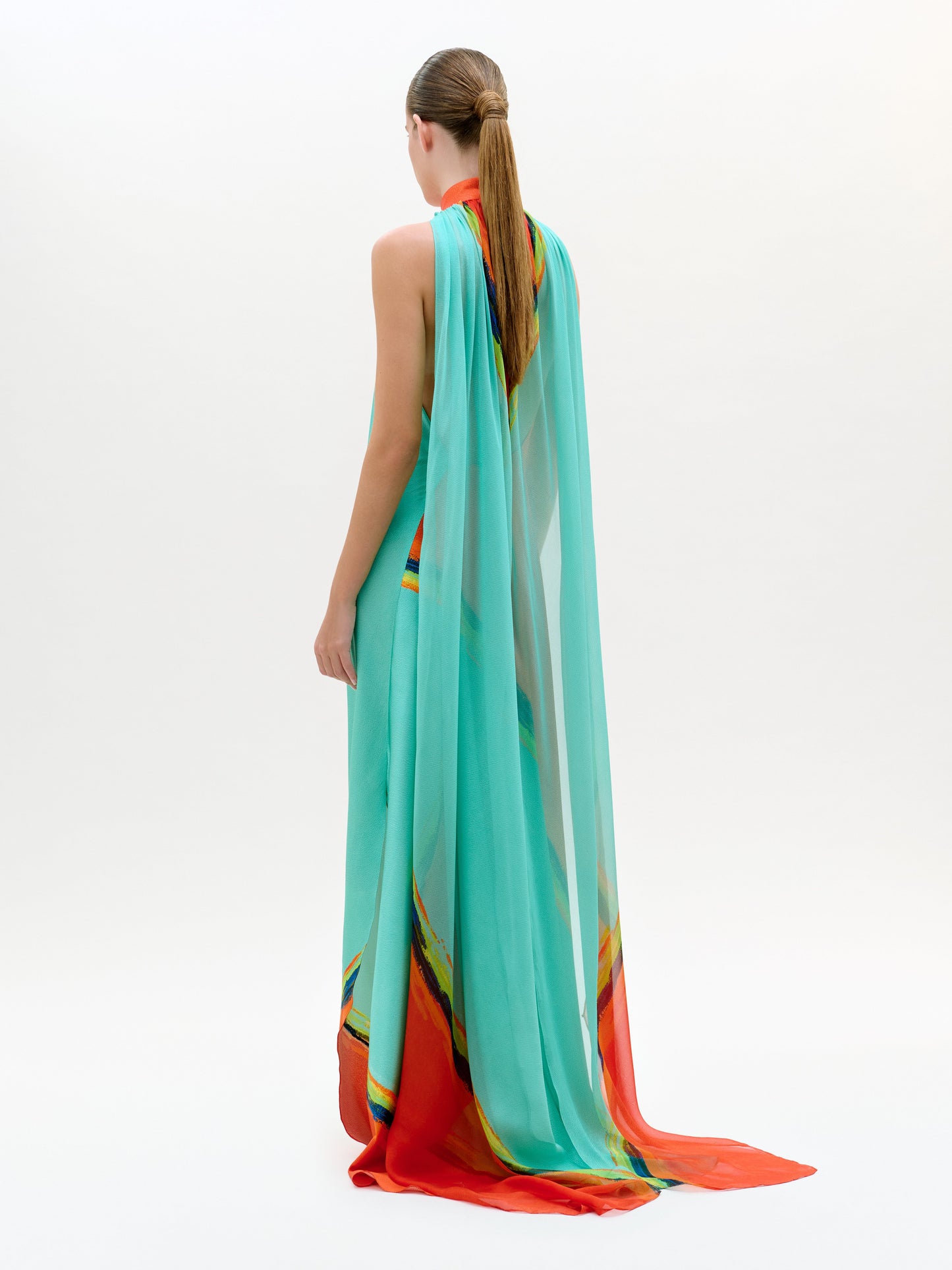 Una persona, de espaldas a la cámara, lleva el vestido Sabina, un maxi largo y vaporoso de color turquesa con detalles en naranja y amarillo y una capa transparente. Lleva el pelo recogido en una elegante coleta sobre un fondo blanco liso.