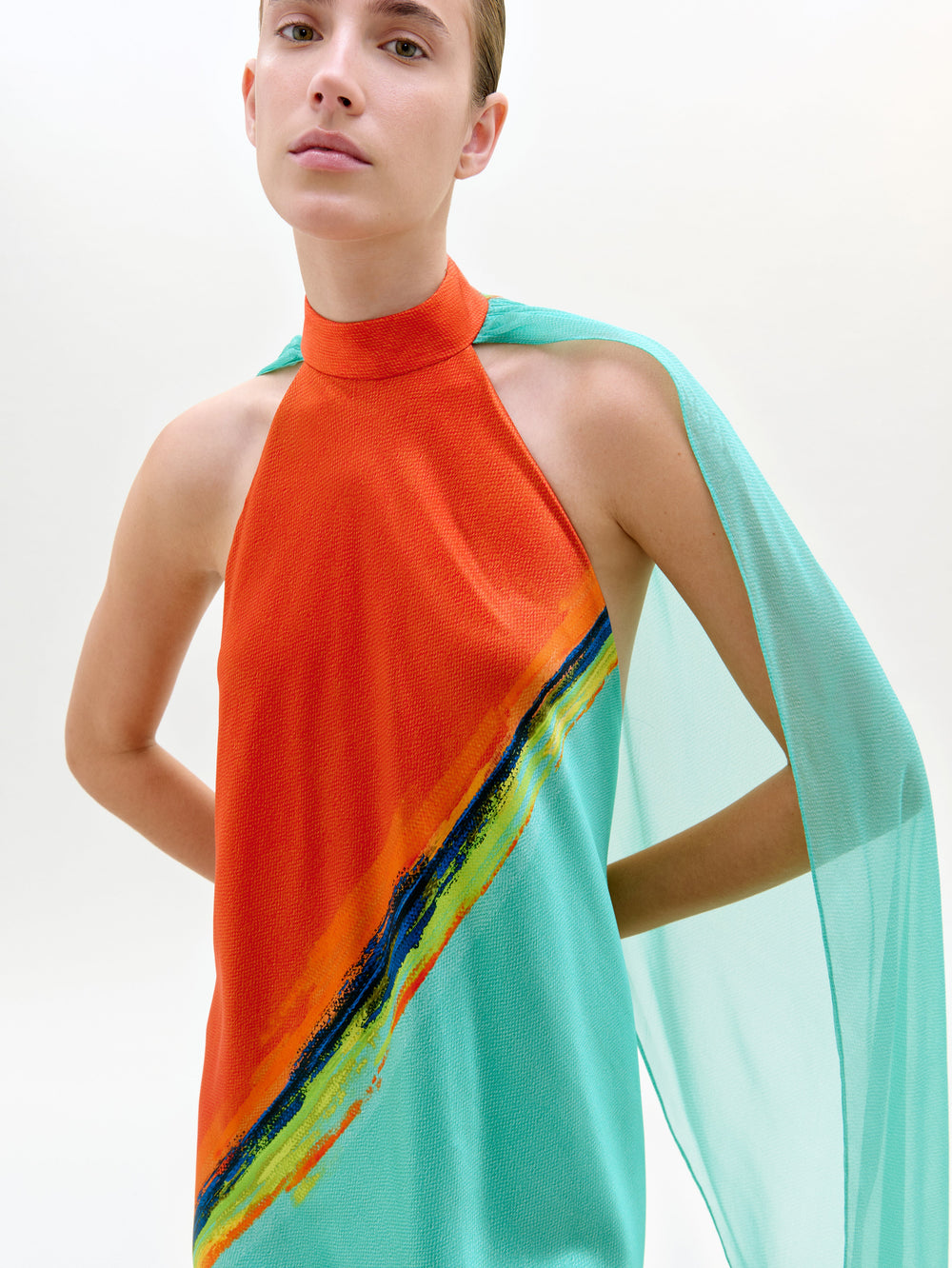 Con el vestido Sabina -un maxi sin mangas con top naranja, tejido italiano turquesa y rayas diagonales multicolores-, una persona se coloca sobre un fondo claro y liso.