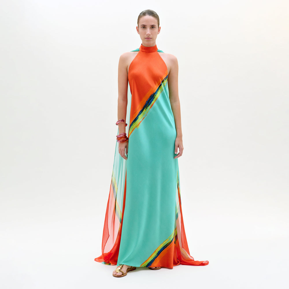 Una persona está de frente con el vestido Sabina, un maxi sin mangas y cuello halter con paneles naranjas y turquesas, rayas diagonales multicolores y secciones transparentes. Lleva el pelo peinado hacia atrás, sandalias y pulseras.