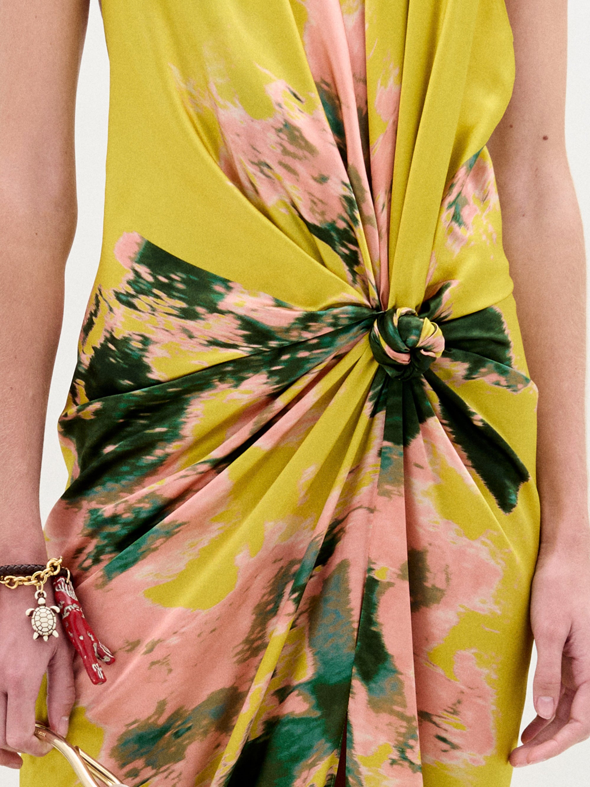 Primer plano del vestido Jazmin, un vestido midi de seda con motivos amarillos, rosas y verdes y cintura de nudo retorcido. La portadora lleva un bolso en la mano izquierda, adornado con pulseras doradas y un colgante rojo.