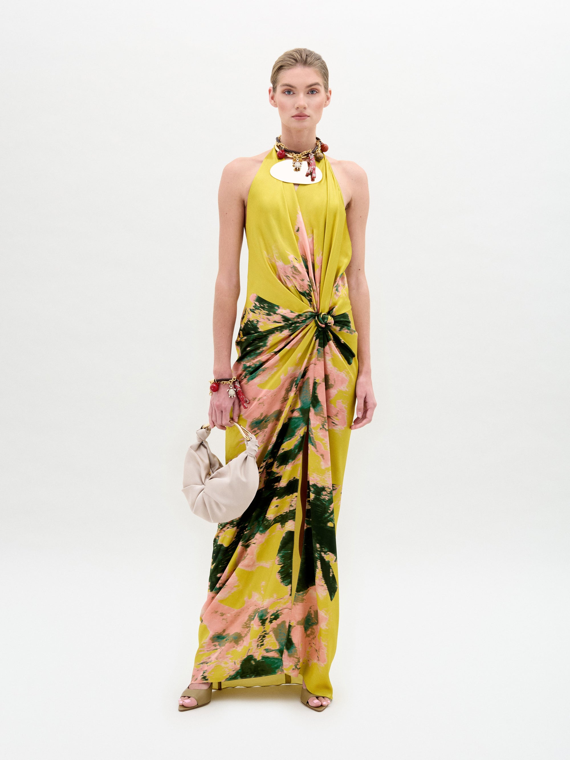 Una mujer posa sobre un fondo blanco con el Jazmin Dress, un vestido amarillo de largo midi con escote halter y motivos abstractos de color rosa verdoso. Lleva un bolso ligero y accesorios como un collar y pulseras.