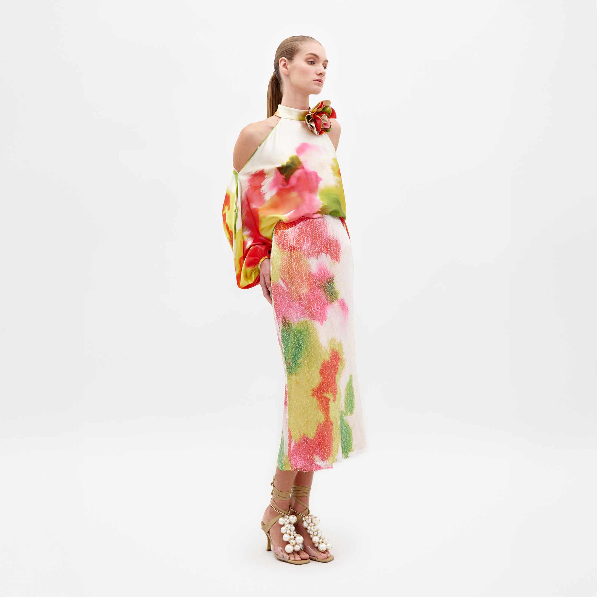 Una mujer está de pie sobre un fondo blanco vistiendo la Falda Sarla, una colorida falda midi con estampado floral hecha de textil italiano, combinada con un top halter a juego y sandalias adornadas con perlas color canela.