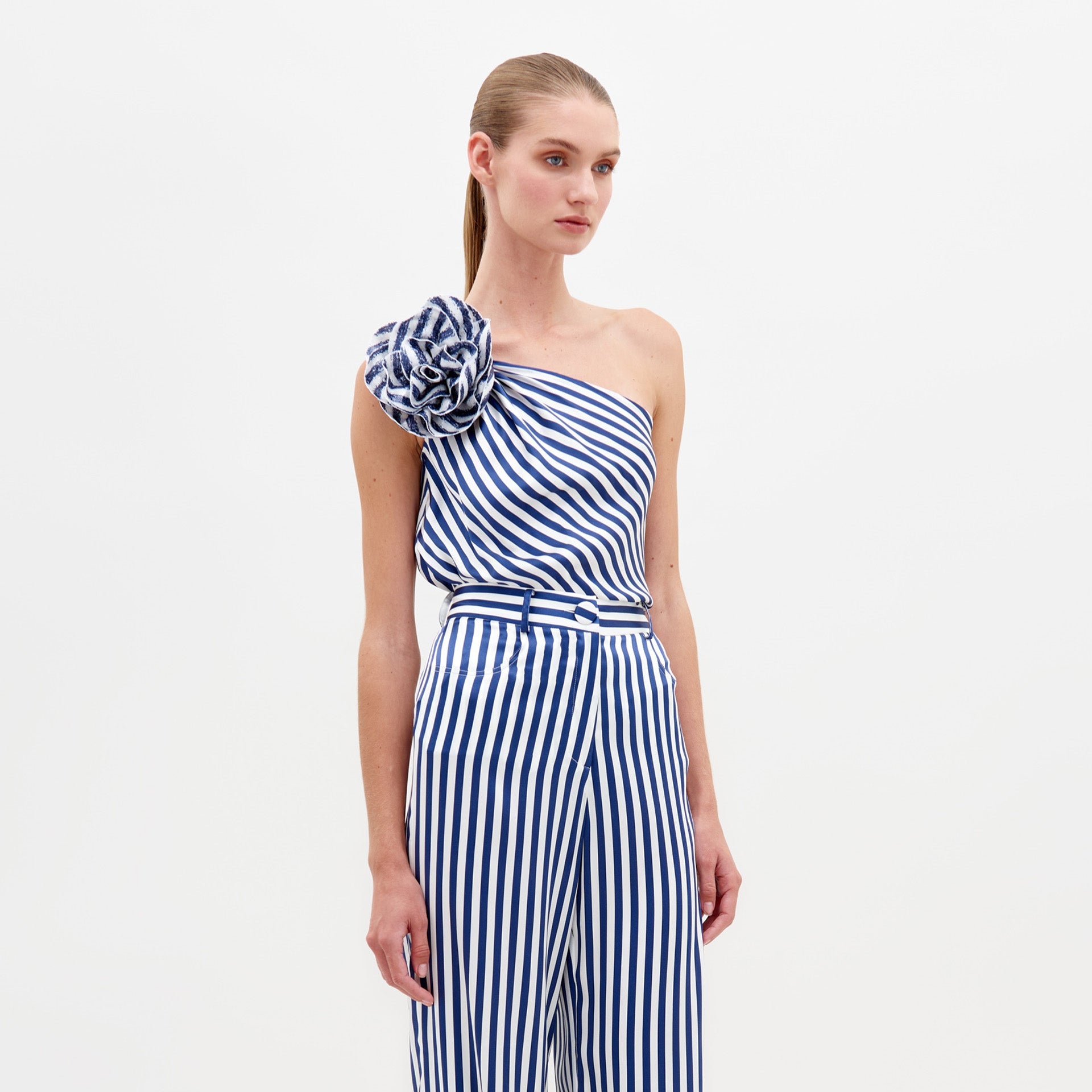 Una mujer viste la Blusa Sebastiana, un top de un solo hombro a rayas azules y blancas con un gran motivo floral de tela en el hombro, sobre un fondo blanco liso.
