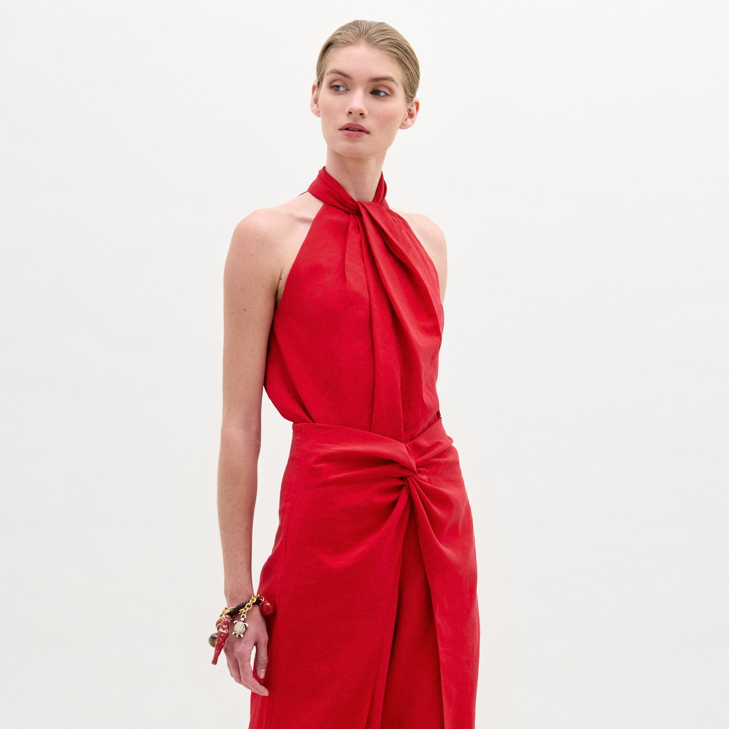 Una persona de pie sobre un fondo blanco liso lleva la blusa roja sin mangas Shela Blouse con escote halter y detalle de cintura anudada, confeccionada en tela italiana, accesorizada con un brazalete grueso, mirando ligeramente hacia un lado.