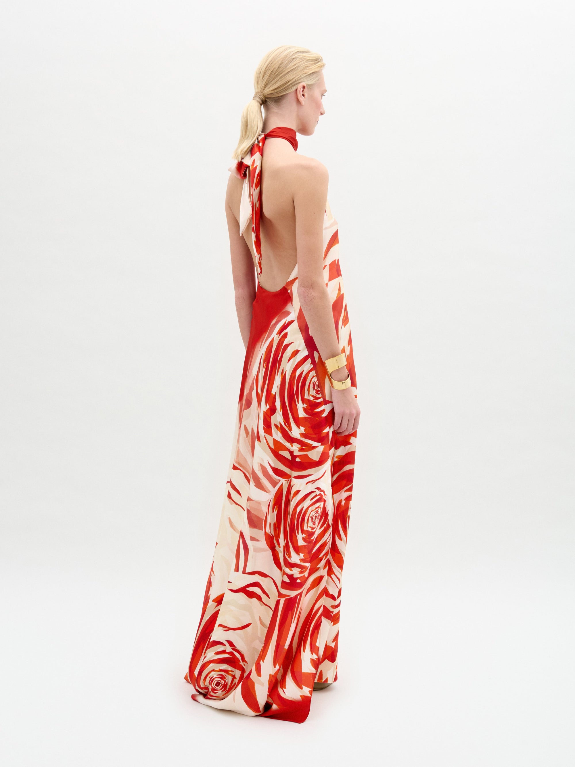 Una persona, de pie sobre un fondo blanco liso, lleva el Sherry Dress, un maxivestido de seda con escote halter, espalda abierta y estampado floral rojo y crema. Lleva el pelo rubio recogido hacia atrás.