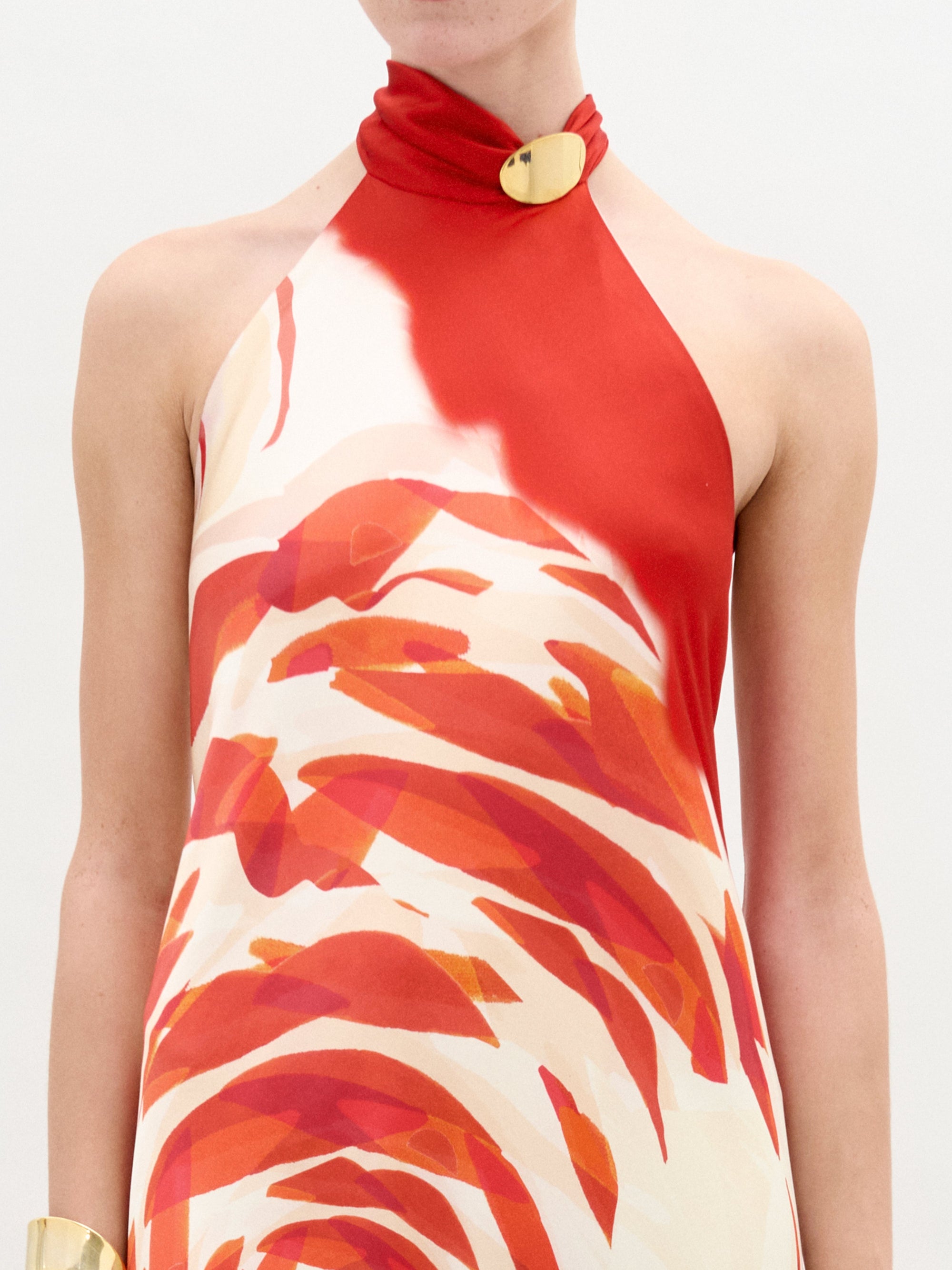 Una persona lleva el vestido Sherry: un maxi de seda sin mangas con un estampado abstracto de remolinos rojos y naranjas, escote halter y cierre dorado en el cuello. En la muñeca izquierda lleva un brazalete ancho dorado. El fondo es blanco liso.