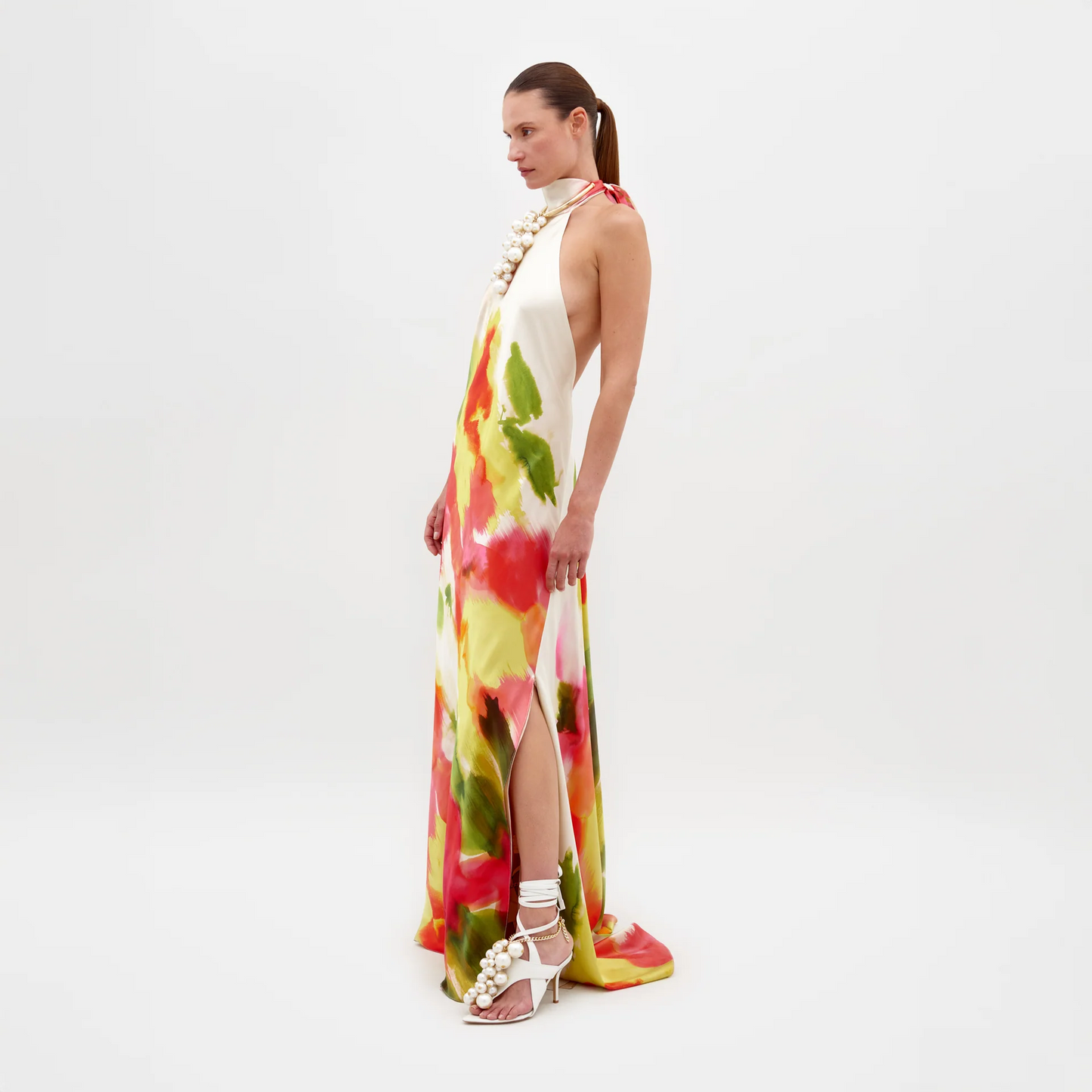 Una mujer de perfil sobre fondo blanco, con el Vestido Sherry de estampado abstracto, tacones blancos y un gran collar de perlas. Lleva el pelo recogido.