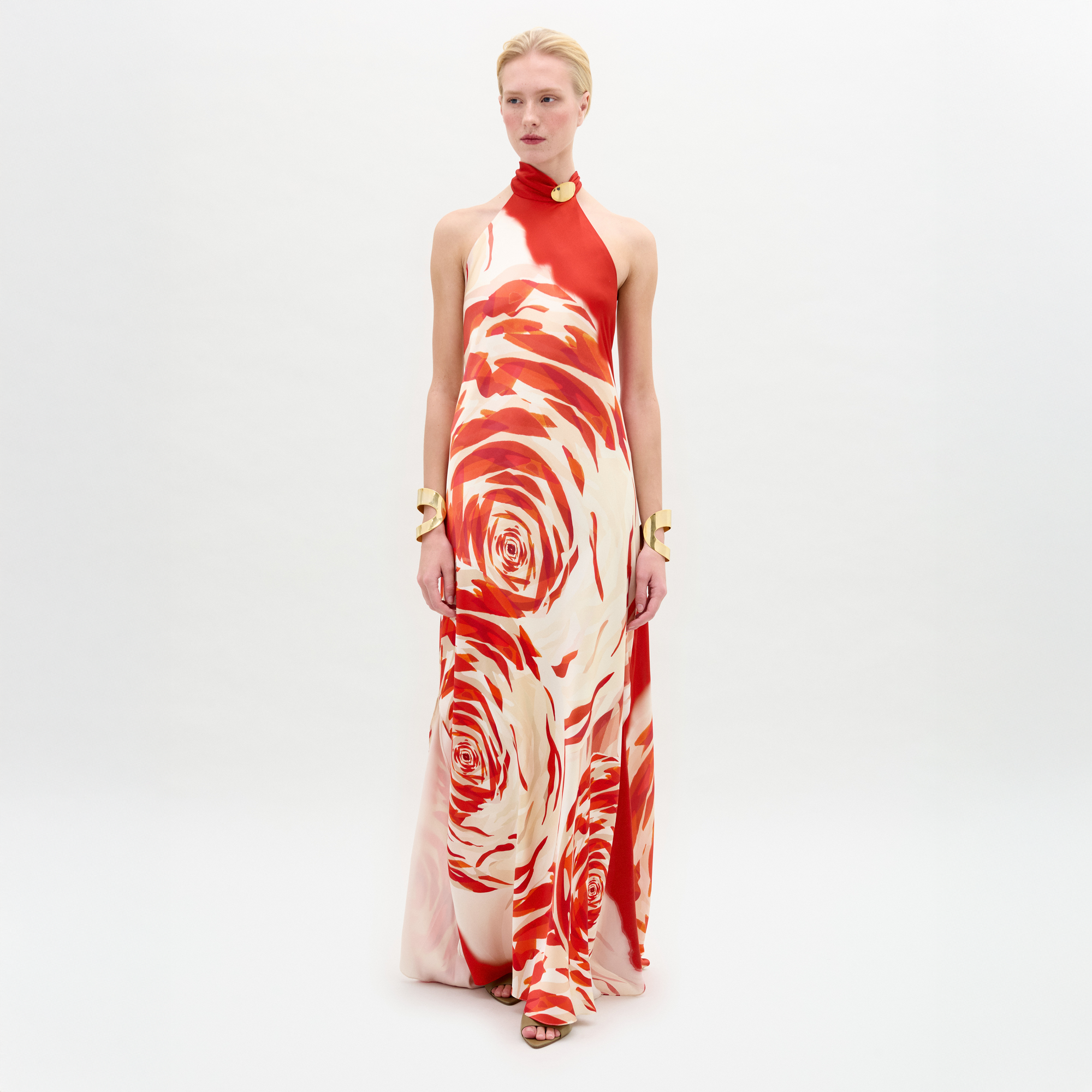 Sobre un fondo blanco liso, una persona luce el vestido Sherry, largo hasta el suelo, con estampado de rosas rojas y blancas, elegante escote halter, brazaletes dorados y zapatos abiertos.