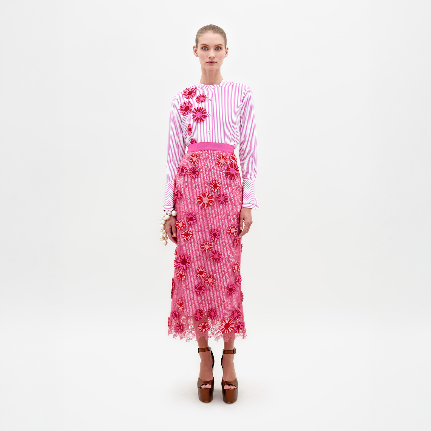 Una mujer aparece de pie sobre un fondo blanco liso con un conjunto a juego, que incluye una blusa blanca con rayas rosas y detalles florales. Su "Falda Uma" luce un diseño floral tridimensional con apliques rosas texturizados. Completa el look con sandalias de plataforma marrones y una pulsera blanca.