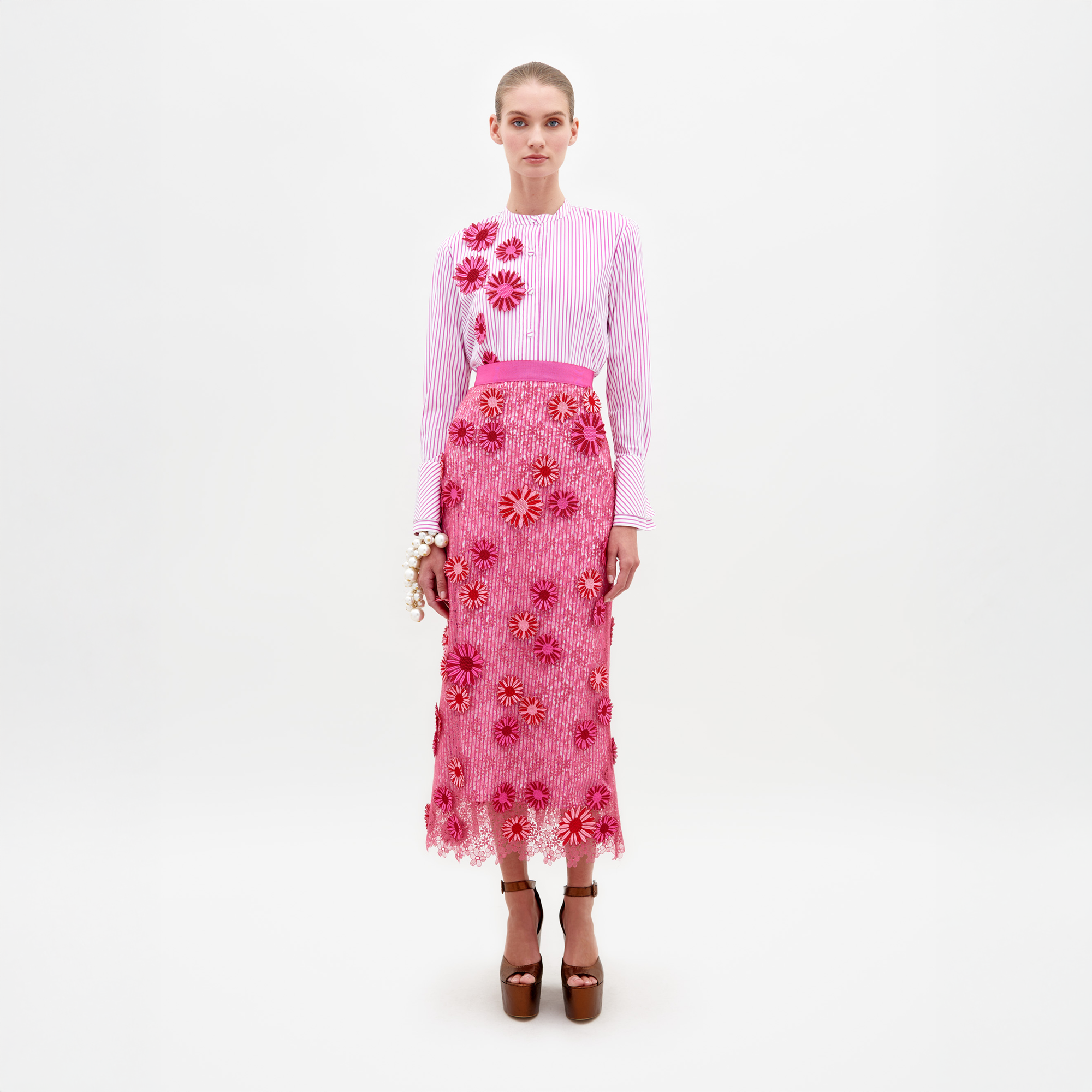 Una mujer aparece de pie sobre un fondo blanco liso con un conjunto a juego, que incluye una blusa blanca con rayas rosas y detalles florales. Su "Falda Uma" luce un diseño floral tridimensional con apliques rosas texturizados. Completa el look con sandalias de plataforma marrones y una pulsera blanca.