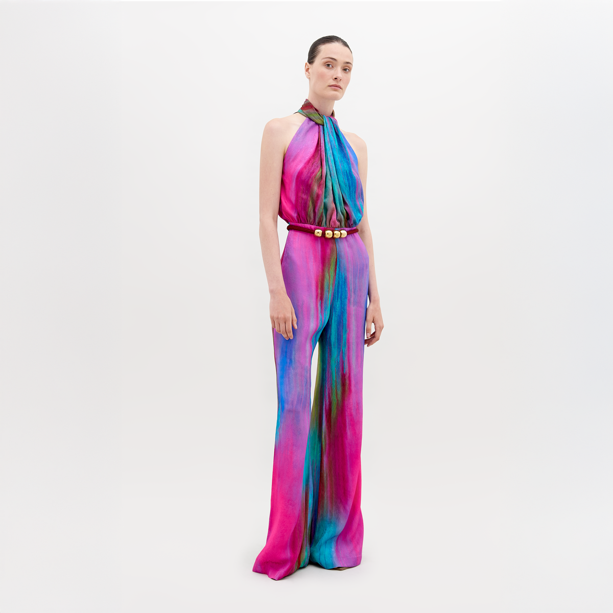 Una persona está parada frente a un fondo blanco con el mono Siv, que presenta tonos vibrantes de rosa, azul y naranja con detalles de cinturón con detalles dorados. El tejido italiano le agrega lujo y el cabello está cuidadosamente recogido.