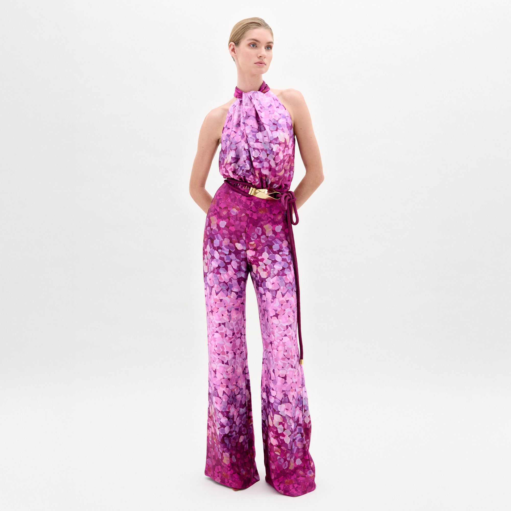 Una mujer posa sobre un fondo blanco con el mono Siv, un mono escote estilo halter con estampado floral rosa y morado y cinturón en la cintura, mirando ligeramente hacia un lado con las manos en las caderas.