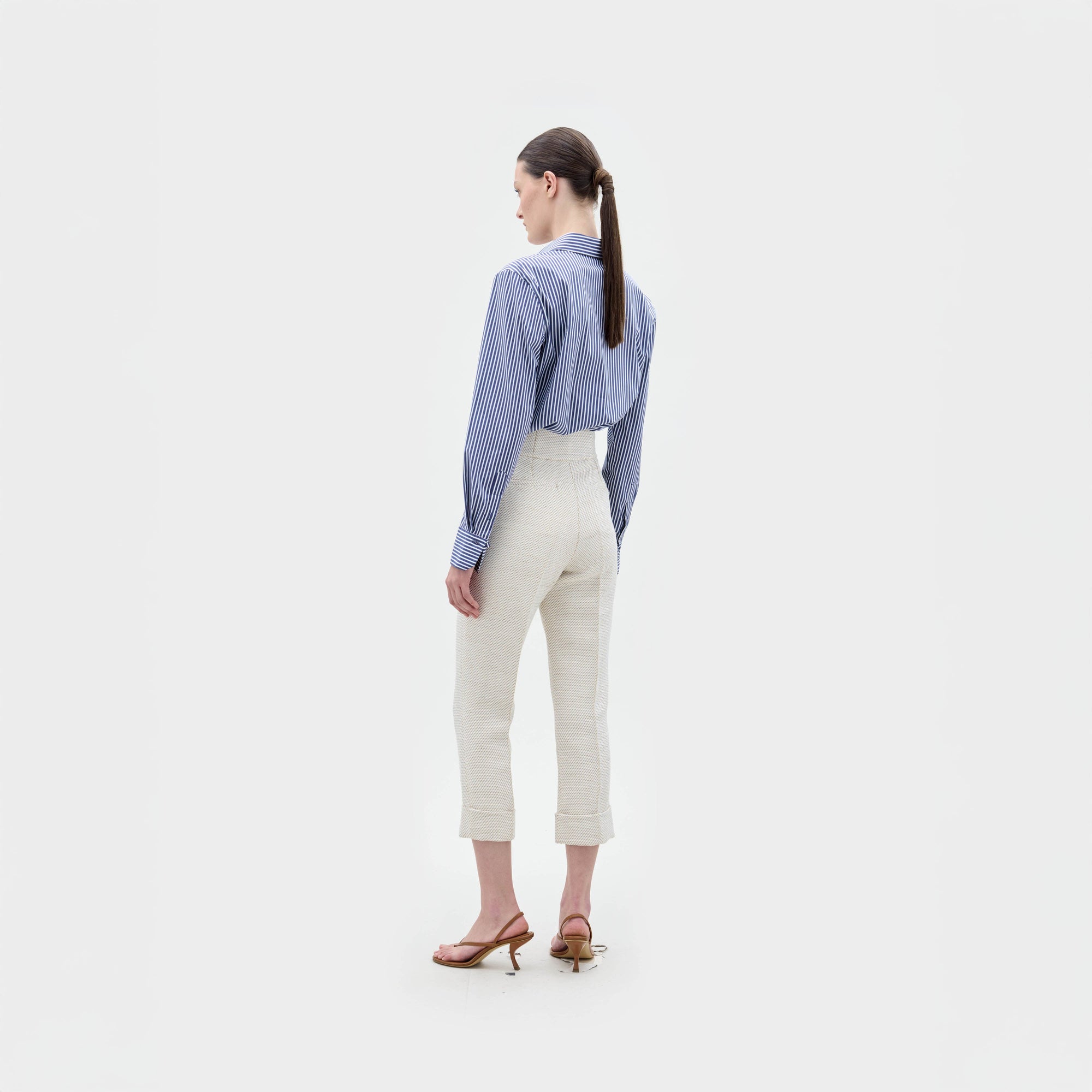 Una mujer mira hacia otro lado, con la blusa Karole a rayas azules, pantalones de talle alto color crema y sandalias de tacón marrones, sobre un fondo blanco liso.