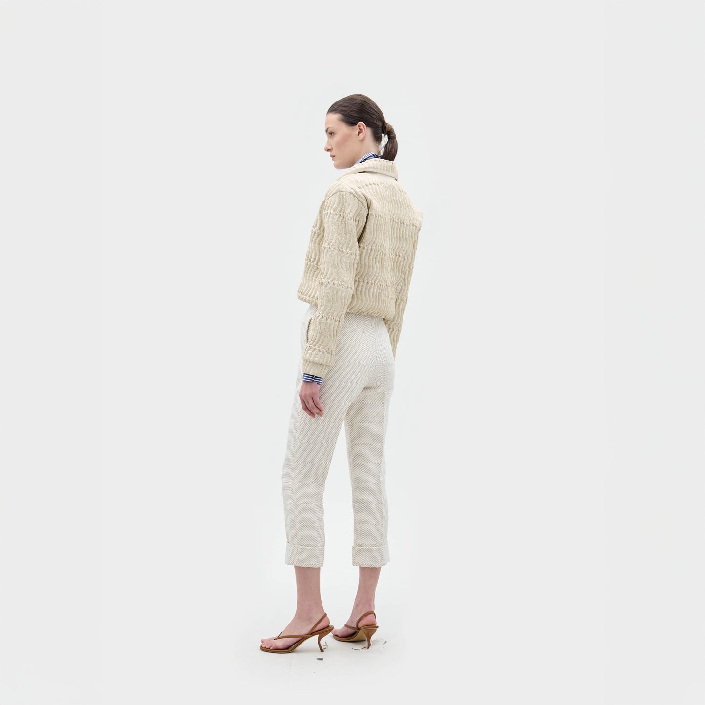 Una persona de pie de lado sobre un fondo blanco liso, con la chaqueta Jaiden, pantalones con puños de color claro, sandalias de tacón y una coleta baja.