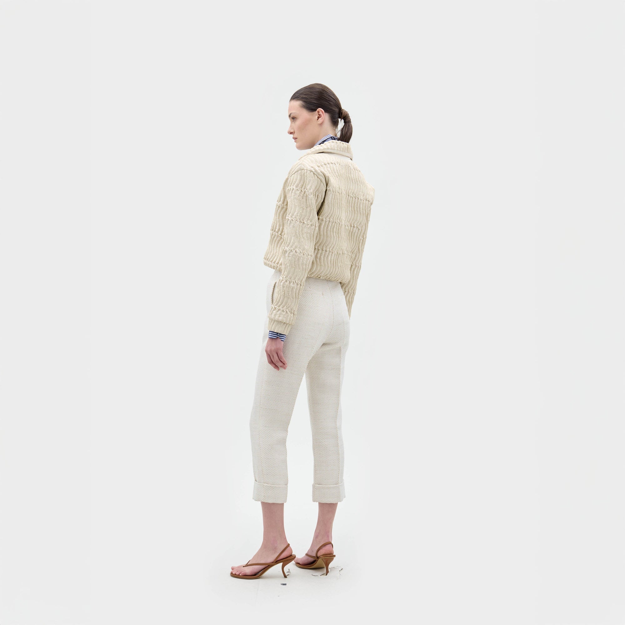 Una persona de pie de lado sobre un fondo blanco liso, con la chaqueta Jaiden, pantalones con puños de color claro, sandalias de tacón y una coleta baja.