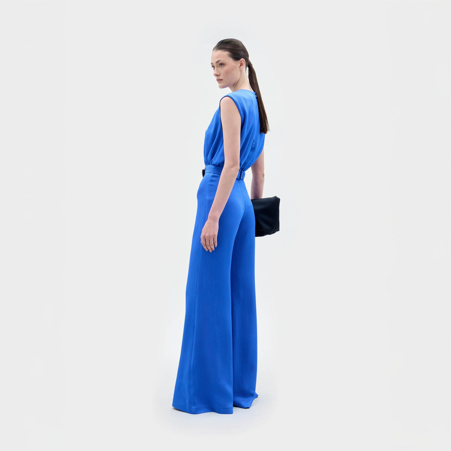 Una mujer posa sobre un fondo blanco vestida con el mono Noel, sin mangas, azul y de pierna ancha. Lleva un bolso de mano negro y mira por encima del hombro. Lleva el pelo recogido en una coleta. Confeccionado en Colombia con tejido italiano.